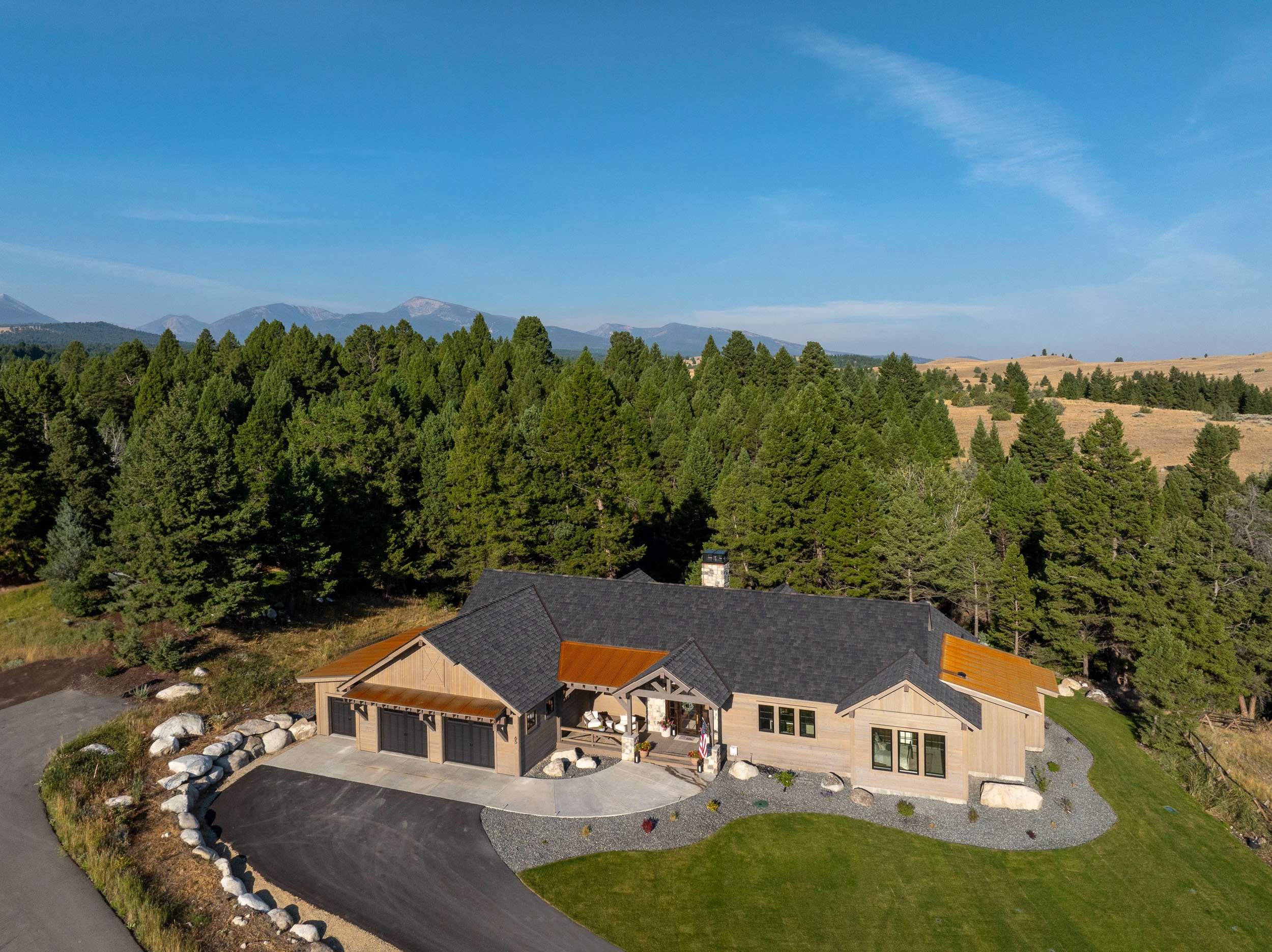 Aerial view of a modern house with a dark roof, wooden exterior, and large windows, set in a green landscape with a driveway, surrounded by trees and mountains in the background. Custom Home built in Montana by Edgell Building.