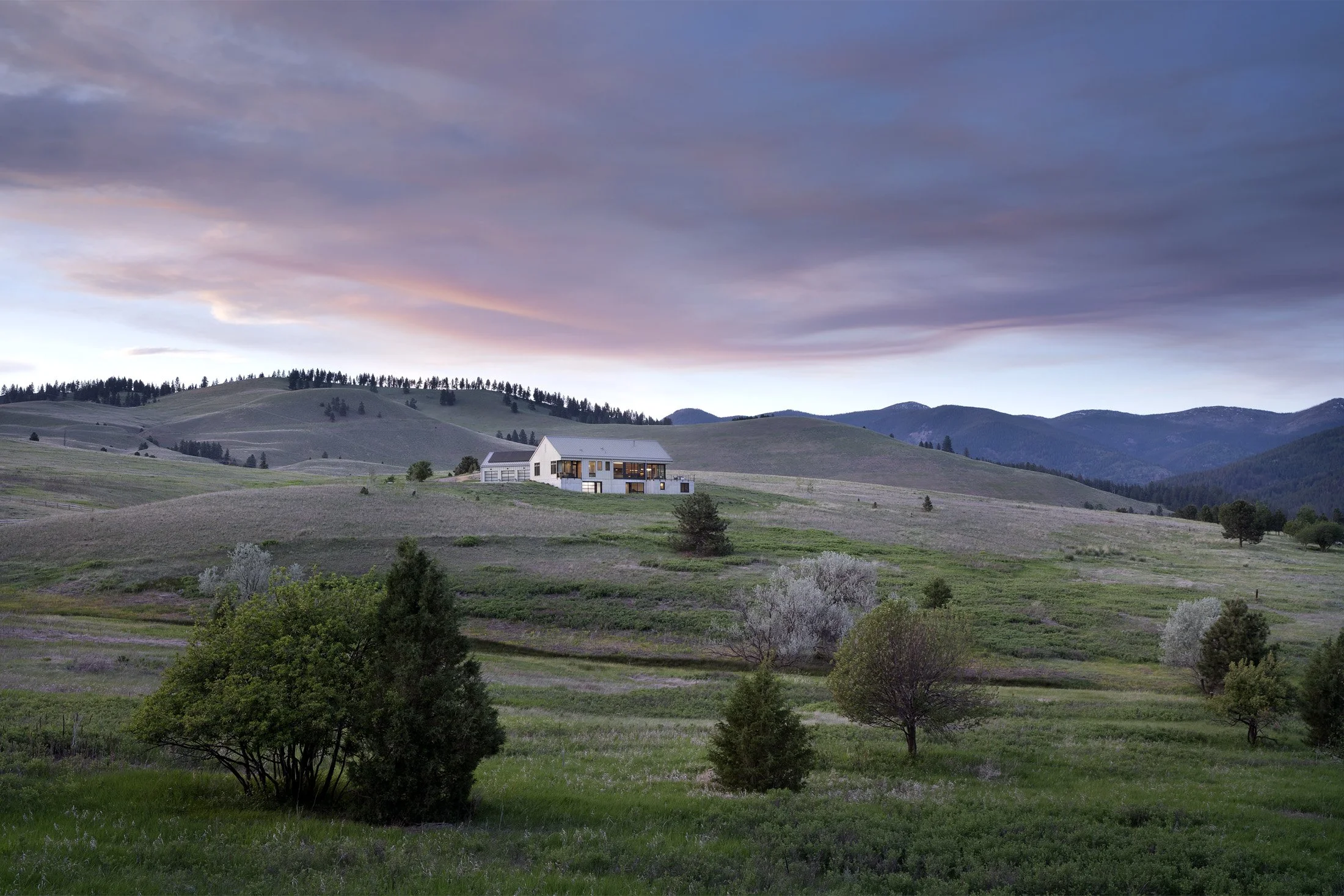 A house on a hilltop surrounded by rolling green fields and scattered trees, with a mountain range in the distance and a colorful sky at sunset. Custom Home built in Montana by Edgell Building.