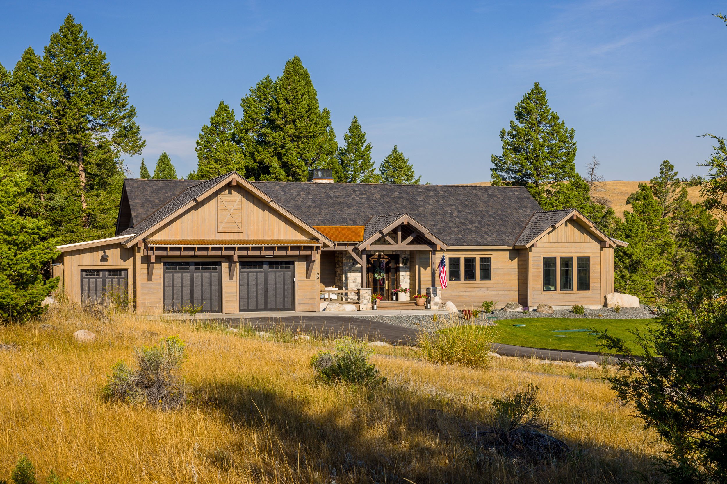 Front of a modern house with a stone and wood exterior, surrounded by greenery and tall trees, under a clear blue sky. Custom Home built in Montana by Edgell Building.