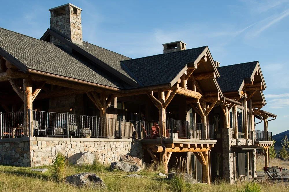 A large mountain-style house with stone and wooden exterior, multiple balconies, and a sloped roof, situated on a grassy landscape with rocks and furniture visible outside. Custom Home built in Montana by Edgell Building.