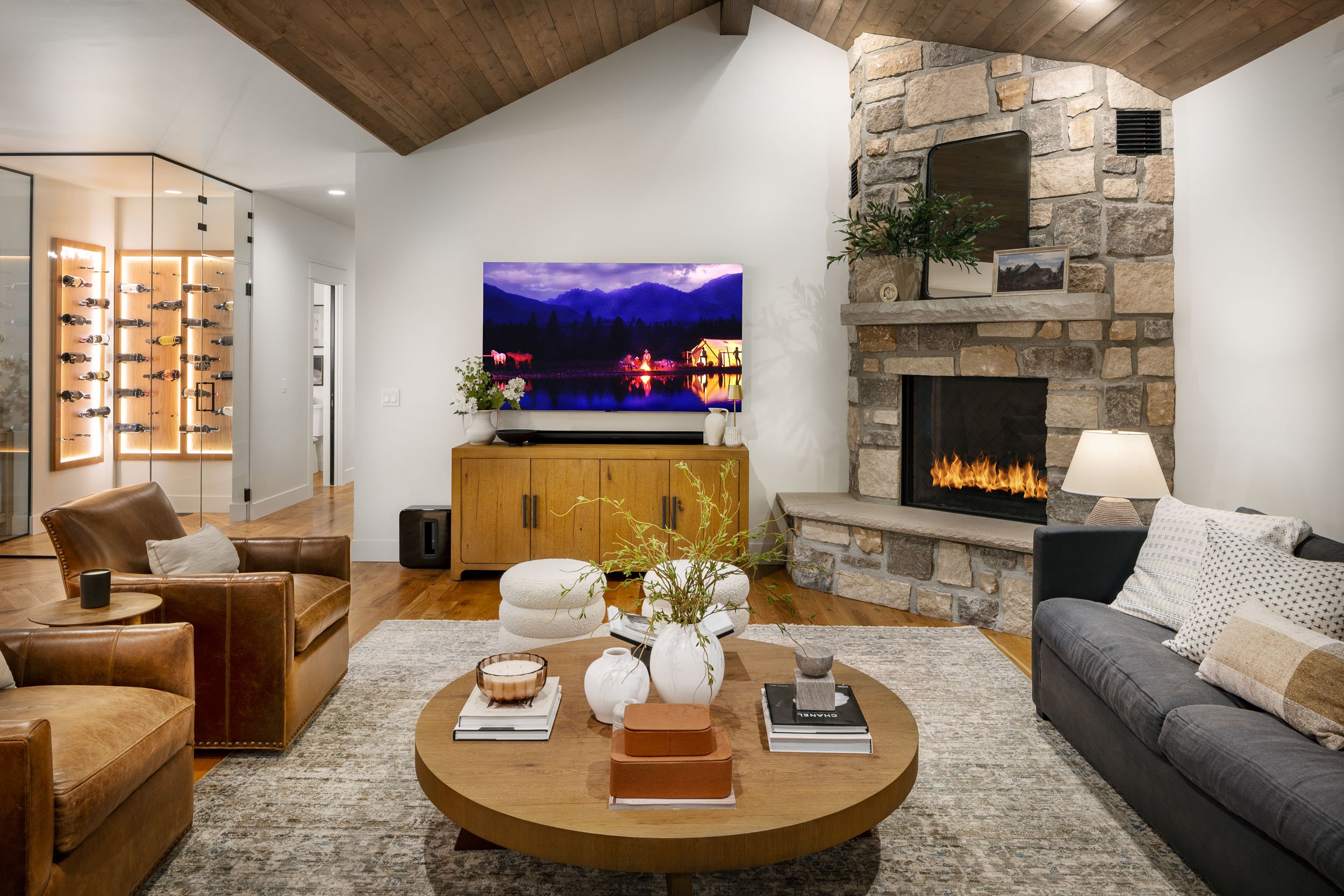 Living room with a stone fireplace, a flat-screen TV on a wooden console, and leather and fabric sofas. A round wooden coffee table with vases, books, and decorative items is in the center. A wall of wine racks and a glass door are visible in the bac