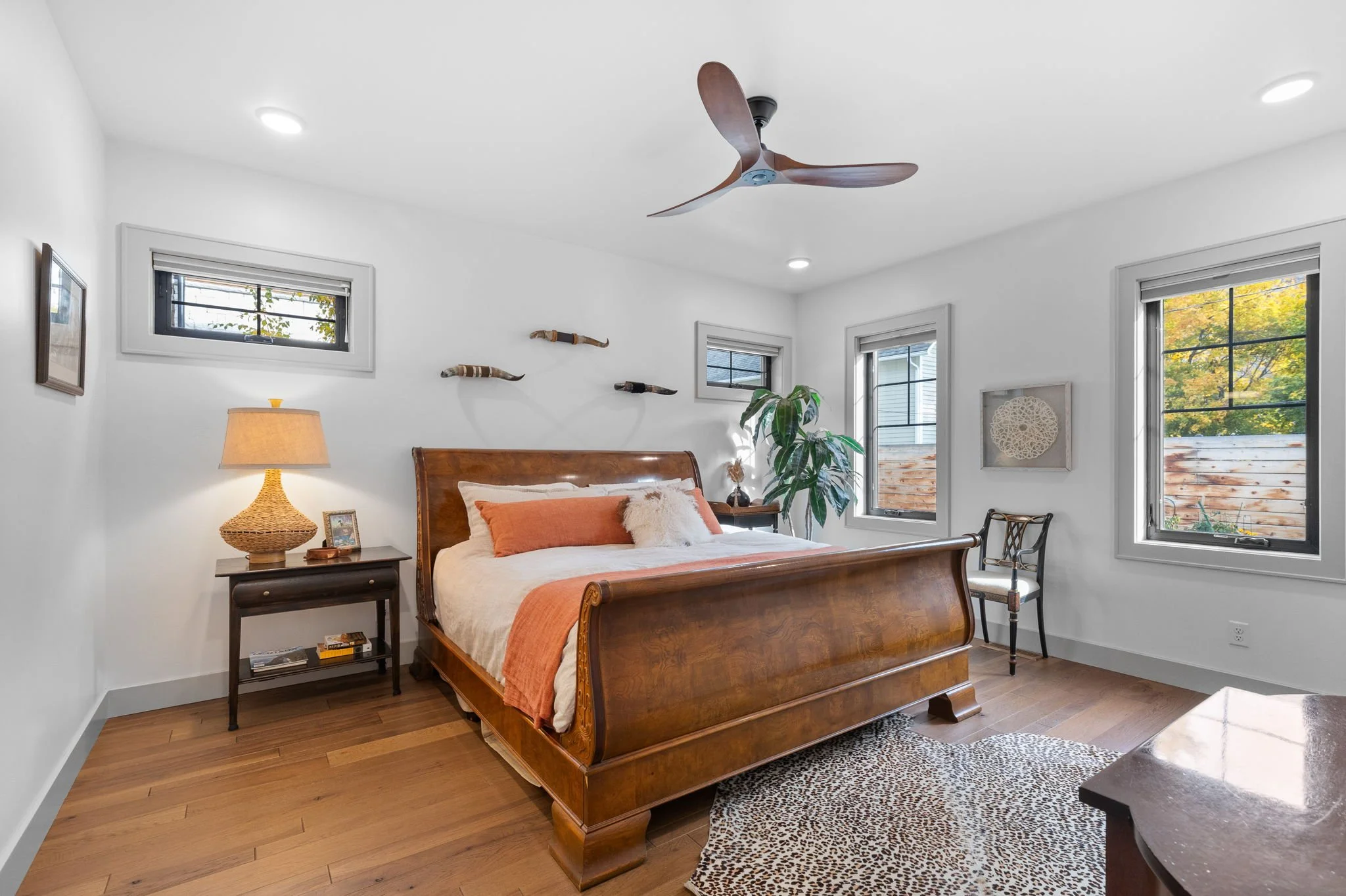 A bedroom with a wooden bed, beige and orange pillows, side table with a lamp, a plant, and three small windows, with a ceiling fan and hardwood floor. Custom Home built in Montana by Edgell Building.