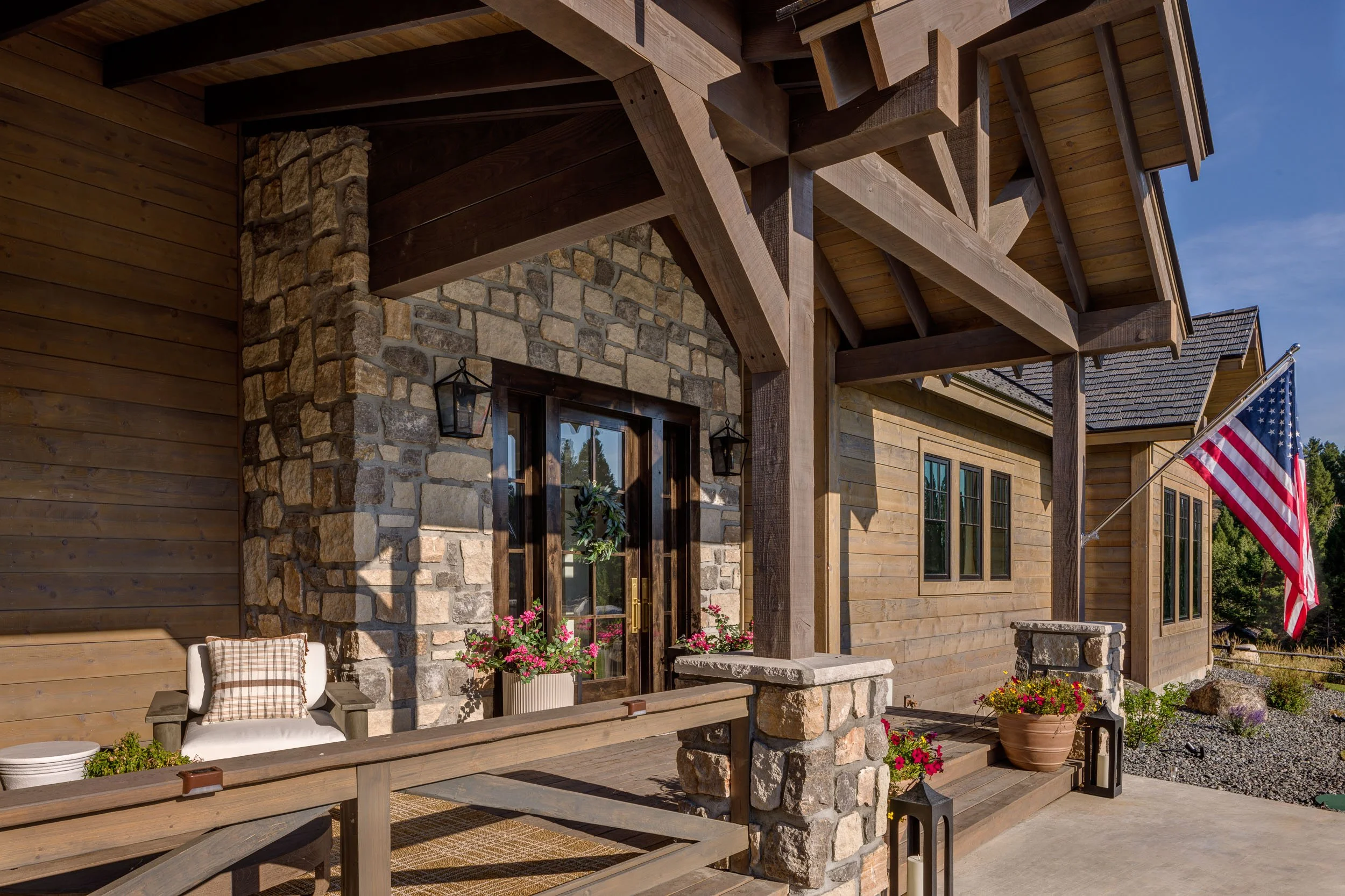 Front porch of a wooden house with stone accents, an American flag, potted flowers, a bench with a pillow, and lanterns. Custom Home built in Montana by Edgell Building.
