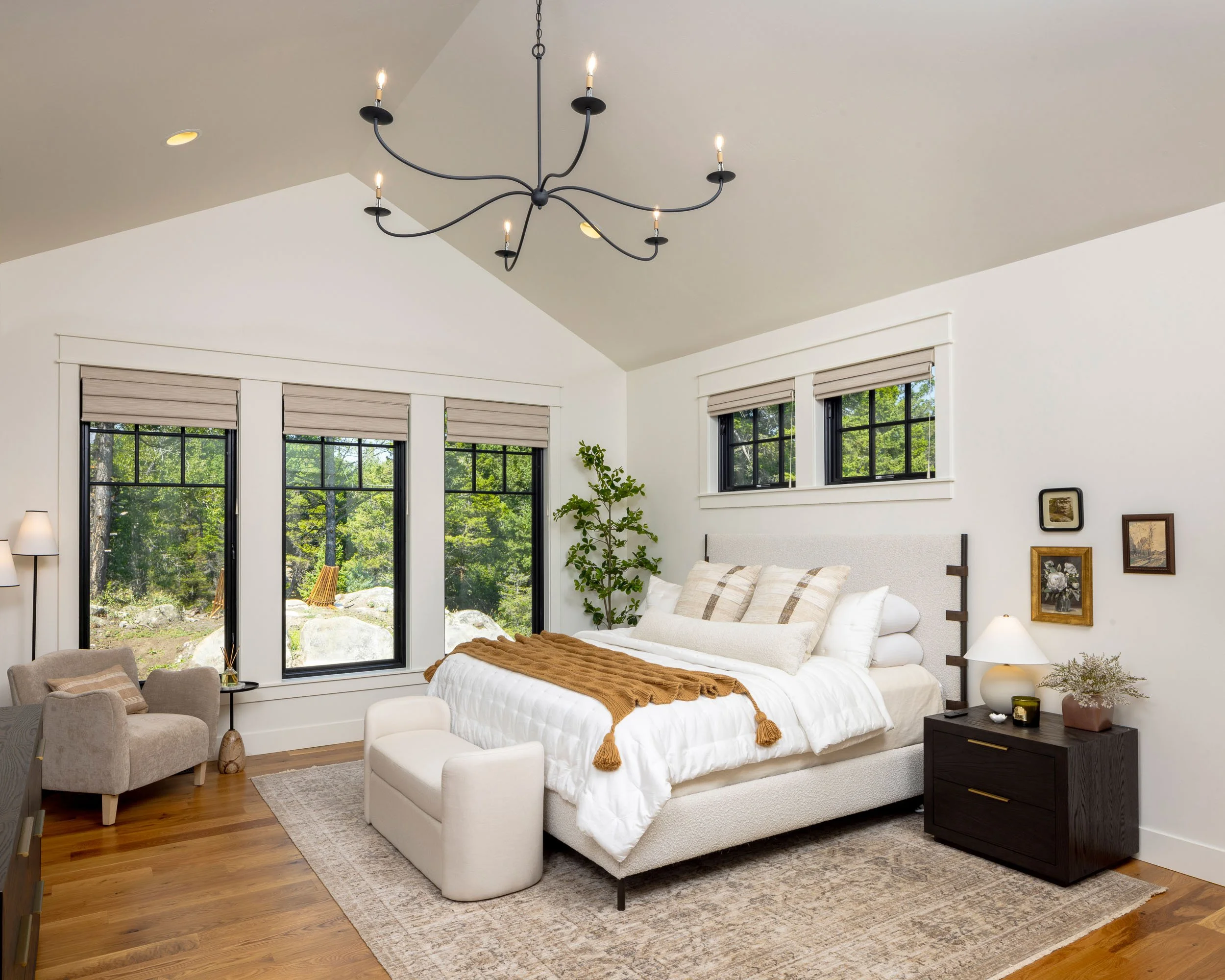 Modern bedroom with a large bed, a black nightstand, a beige armchair, and windows showing greenery outside. Custom Home built in Montana by Edgell Building.