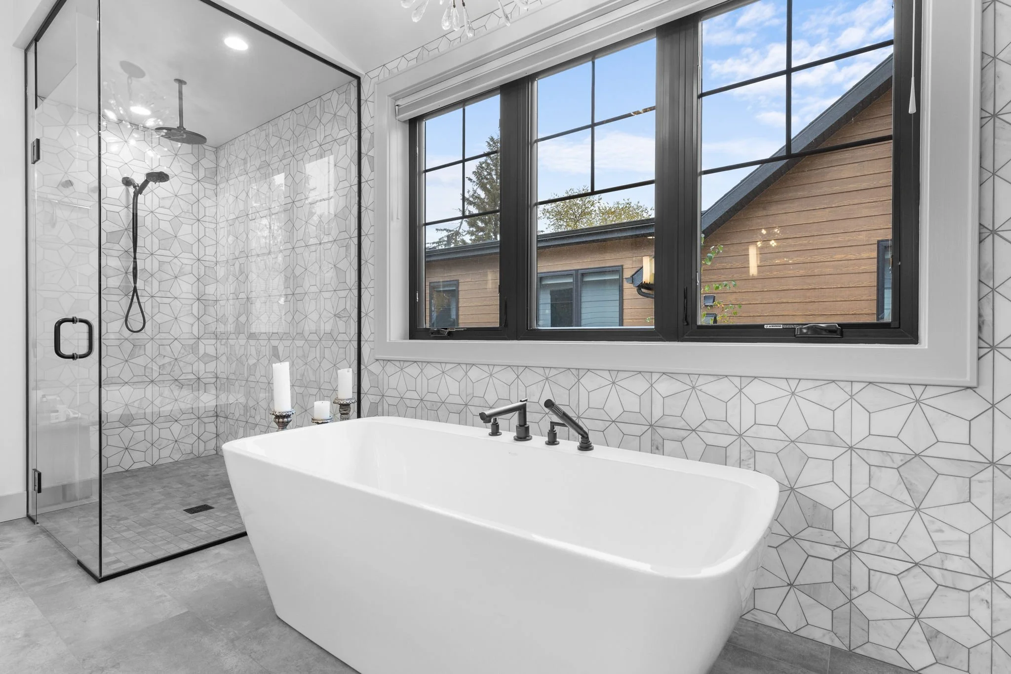 Modern bathroom with a white bathtub, a glass-enclosed shower with geometric tile walls, and large black-framed windows showing a view of neighboring houses and trees. Custom Home built in Montana by Edgell Building.