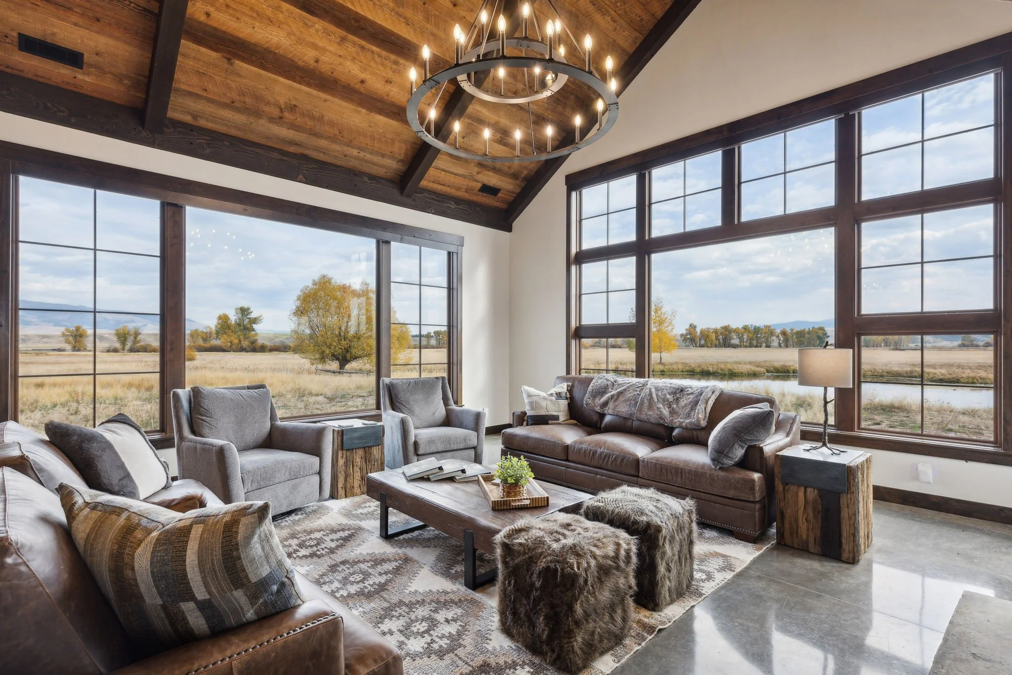 Living room with large windows overlooking a fall landscape, featuring a chandelier, leather and fabric sofas, a patterned rug, and rustic decor. Custom Home built in Montana by Edgell Building.