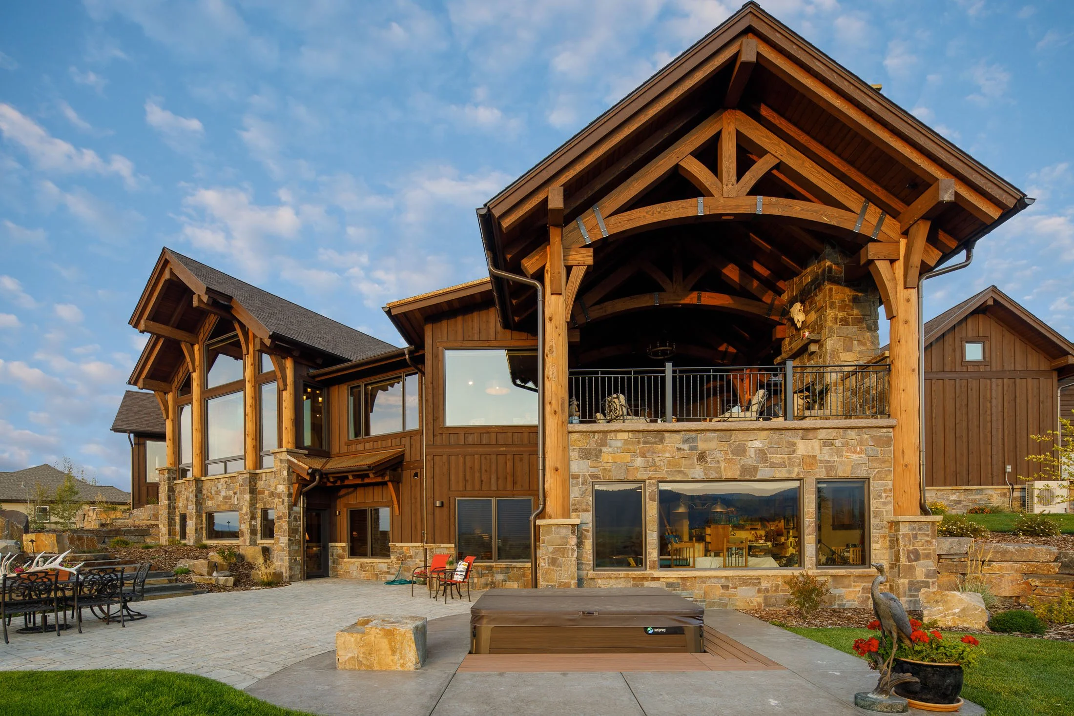 Large house with a stone and wood exterior, multiple large windows, a covered balcony with chairs, and an outdoor hot tub, set against a blue sky with scattered clouds. Custom Home built in Montana by Edgell Building.