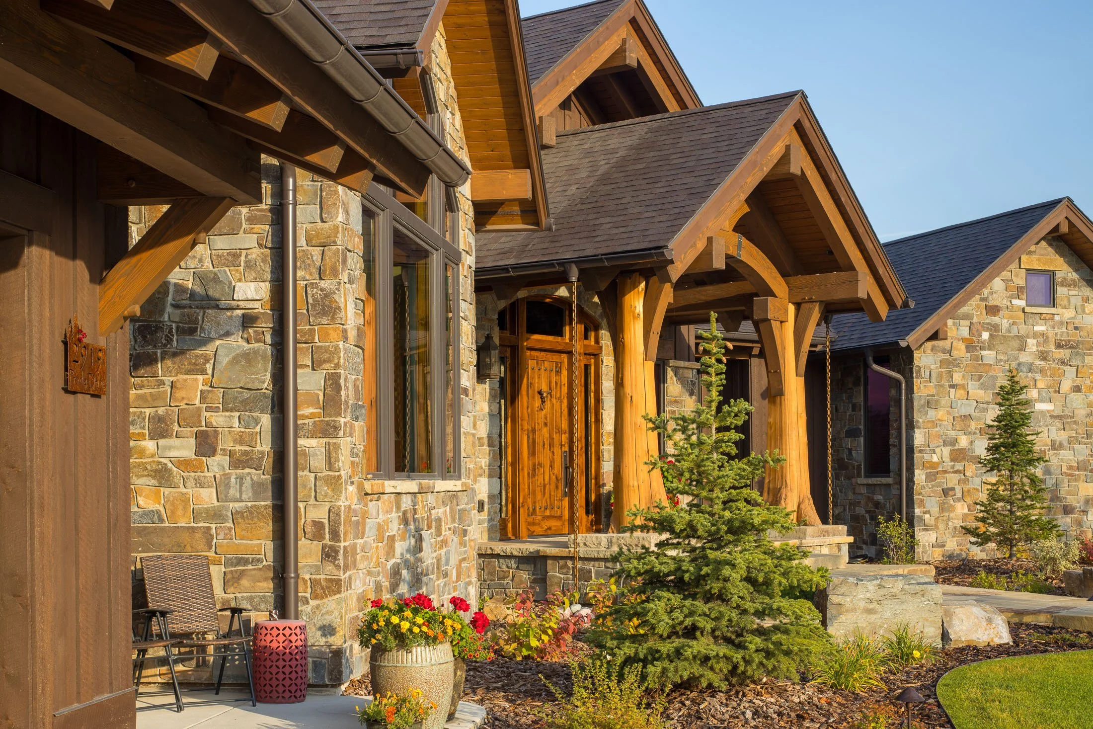 A house with stone and wood exterior, front porch with wooden beams, and small evergreen trees in the landscaped yard. Custom Home built in Montana by Edgell Building.