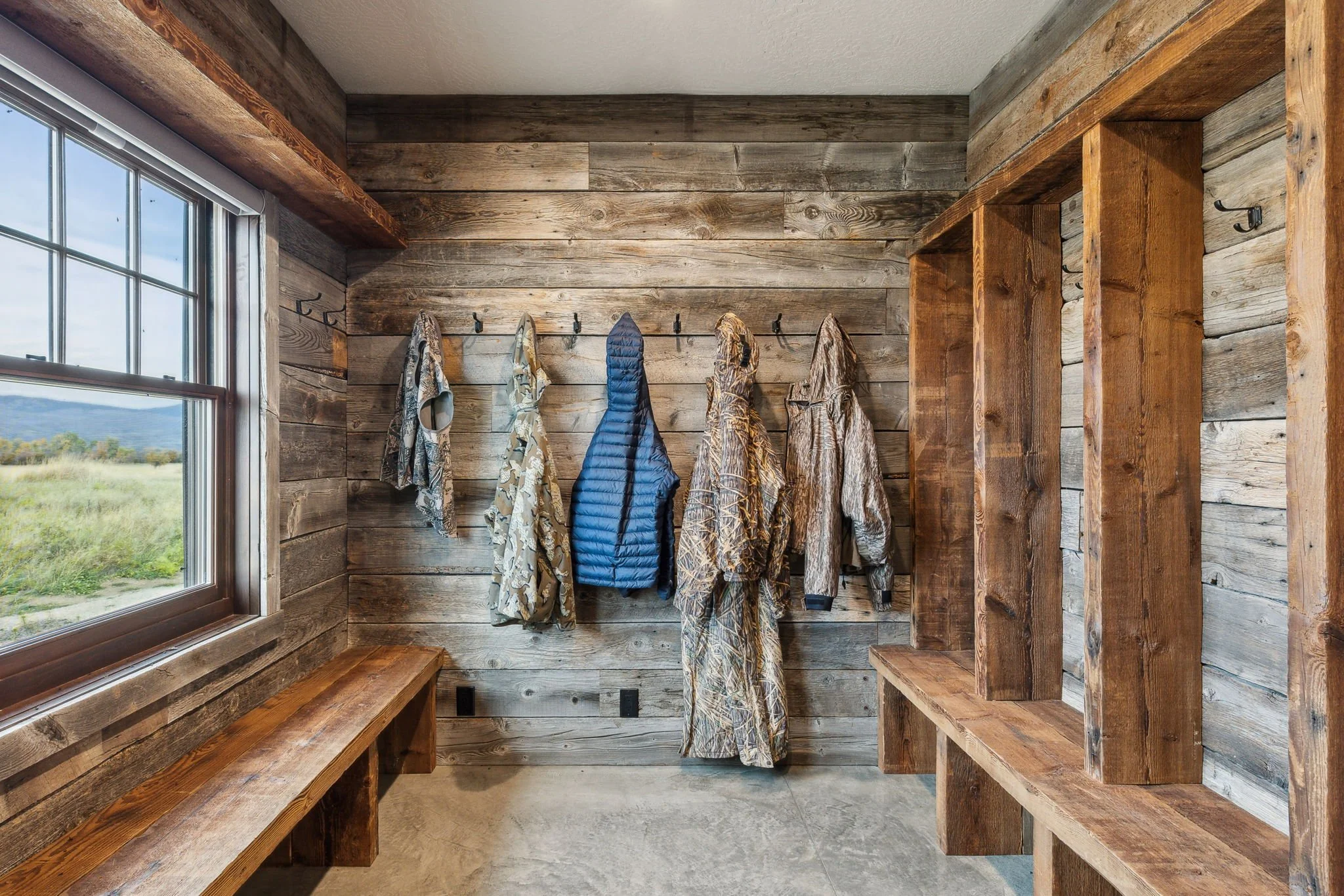 Rustic mudroom with wooden walls, hooks holding camouflage clothing and a blue vest, a bench under a window showing a view of a grassy landscape. Custom Home built in Montana by Edgell Building.