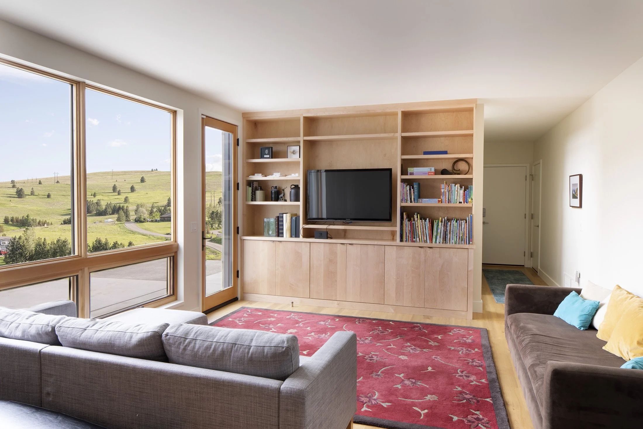 Living room with large windows overlooking a hilly landscape, light wood built-in entertainment center with a TV, bookshelves with books, and a red area rug, with couches and pillows. Custom Home built in Montana by Edgell Building.