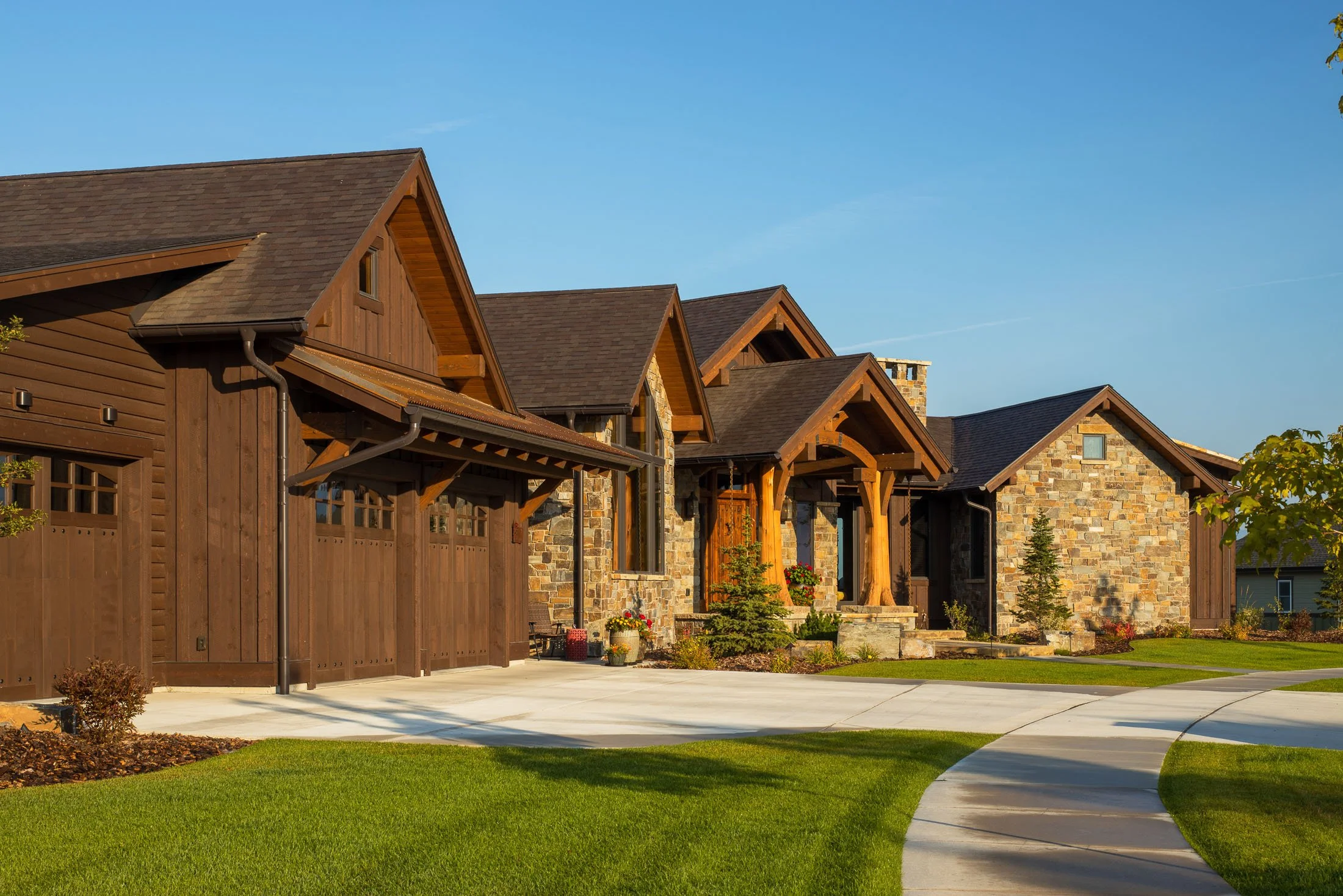 A modern house with a stone and wood exterior, featuring a brown shingled roof, a driveway, and a well-maintained front yard with green grass and small trees. Custom Home built in Montana by Edgell Building.