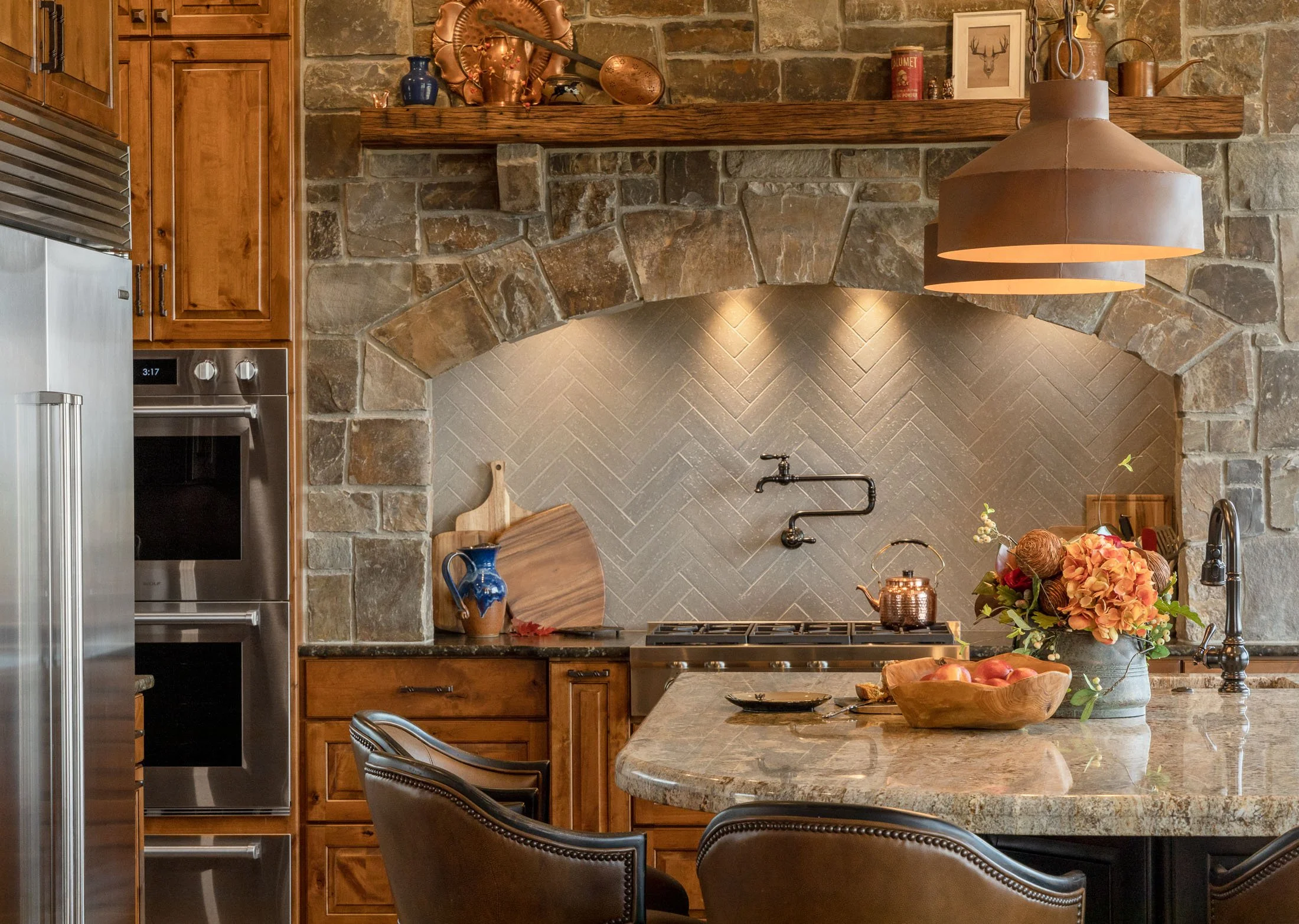 Kitchen with stone and wooden elements, a tiled backsplash, a large kitchen island with a granite countertop, a vase with flowers, and decorative items on a shelf above the stove. Custom Home built in Montana by Edgell Building.