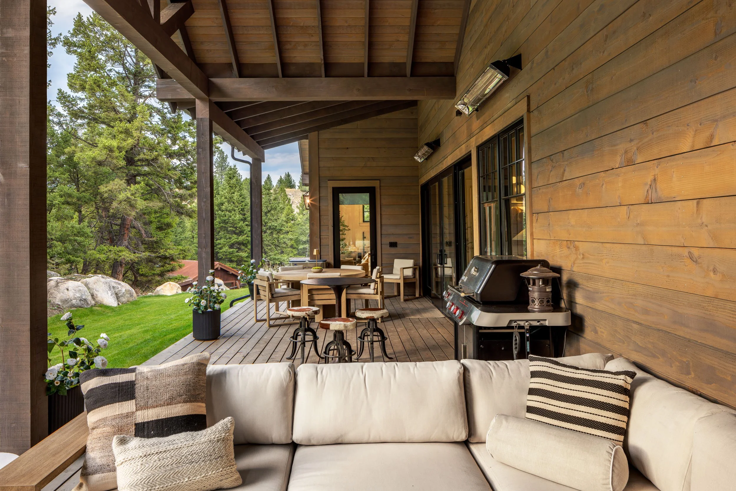 Outdoor covered patio with beige sectional sofa, black and beige pillows, a black grill, and wooden furniture, overlooking a yard with green grass, trees, and large rocks.