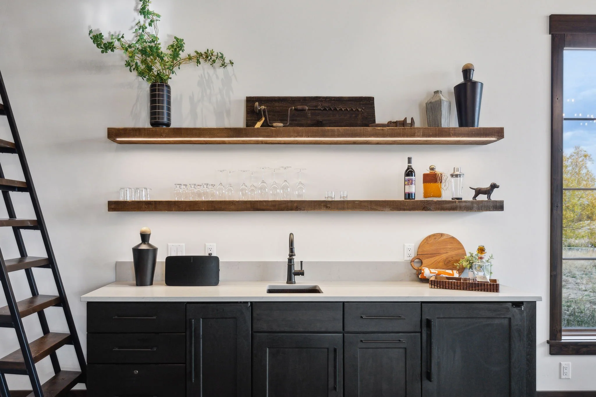 Kitchen with black cabinets, white marble countertop, open wooden shelves with decorative items, window showing outdoor trees. Custom Home built in Montana by Edgell Building.