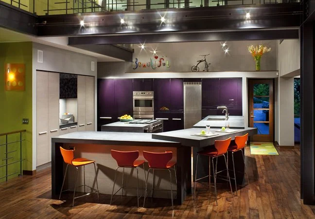Modern kitchen with purple cabinets, a large central island with orange chairs, stainless steel appliances, and decorative wall art and bicycle on the back wall. Custom Home built in Montana by Edgell Building.