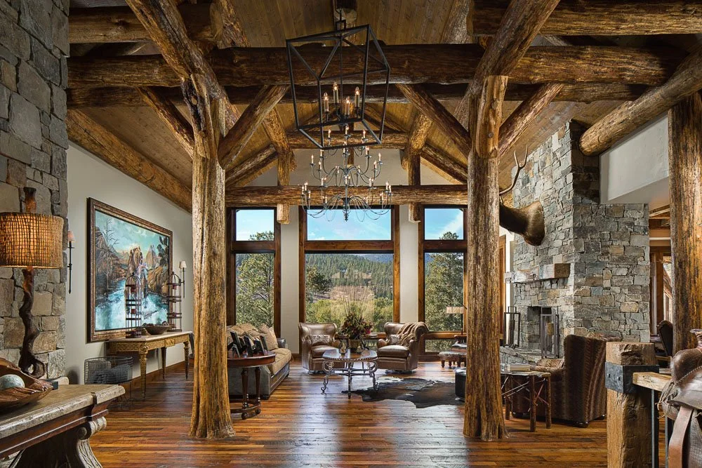Rustic living room with large windows, wooden beams, stone fireplace, leather furniture, and mounted deer head. Custom Home built in Montana by Edgell Building.
