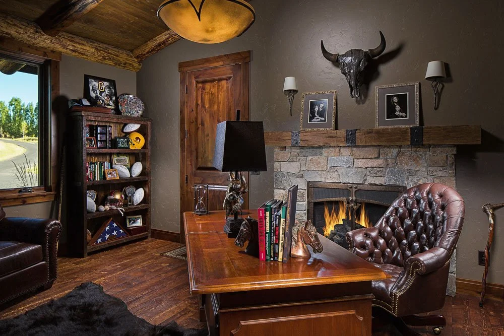 Cozy office with stone fireplace, leather chair, wooden desk, bookshelf with football memorabilia, and rustic decor including animal skull and framed photos. Custom Home built in Montana by Edgell Building.