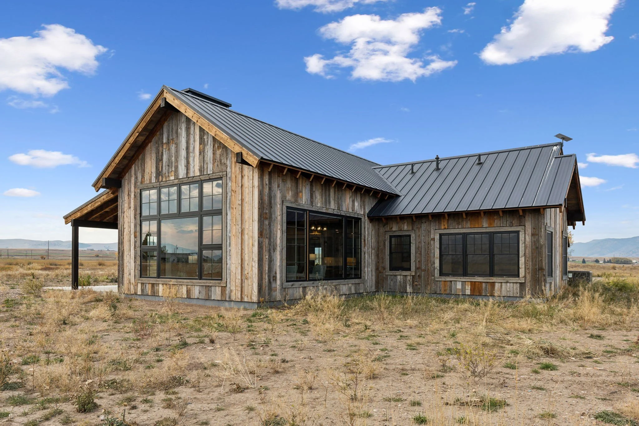 A modern house with wood siding and a metal roof, located in a rural area with dry grass and a clear blue sky with scattered clouds. Custom Home built in Montana by Edgell Building.