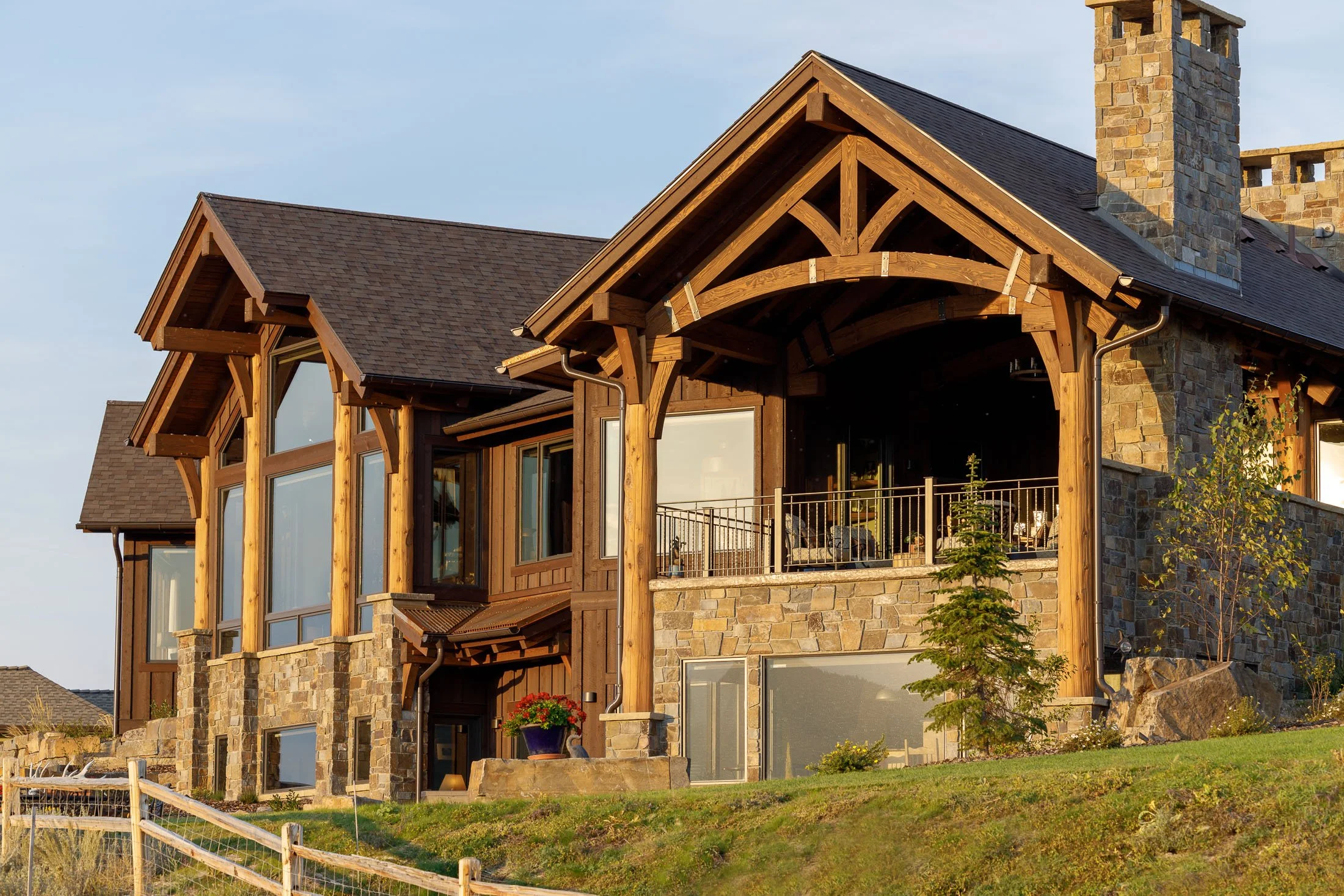 A large modern house with a combination of stone and wood exterior, large windows, a spacious balcony, and a sloped roof, surrounded by a grassy yard with small trees. Custom Home built in Montana by Edgell Building.