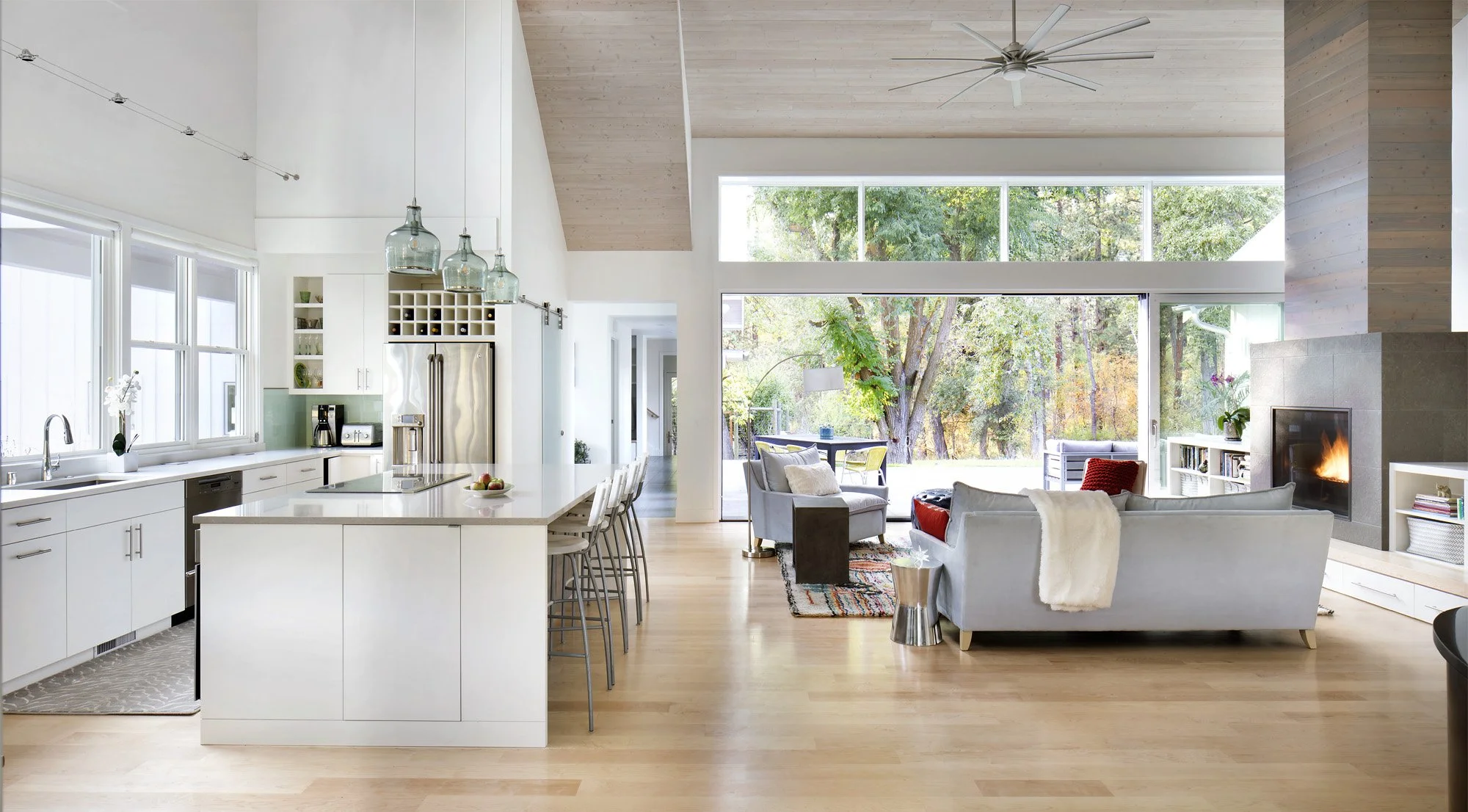 Open-concept living room and kitchen with large windows, white cabinetry, and a neutral color palette, featuring a kitchen island with bar stools, a cozy seating area with sofas and a fireplace. Home Designed and Built By Edgell Building in Montana.
