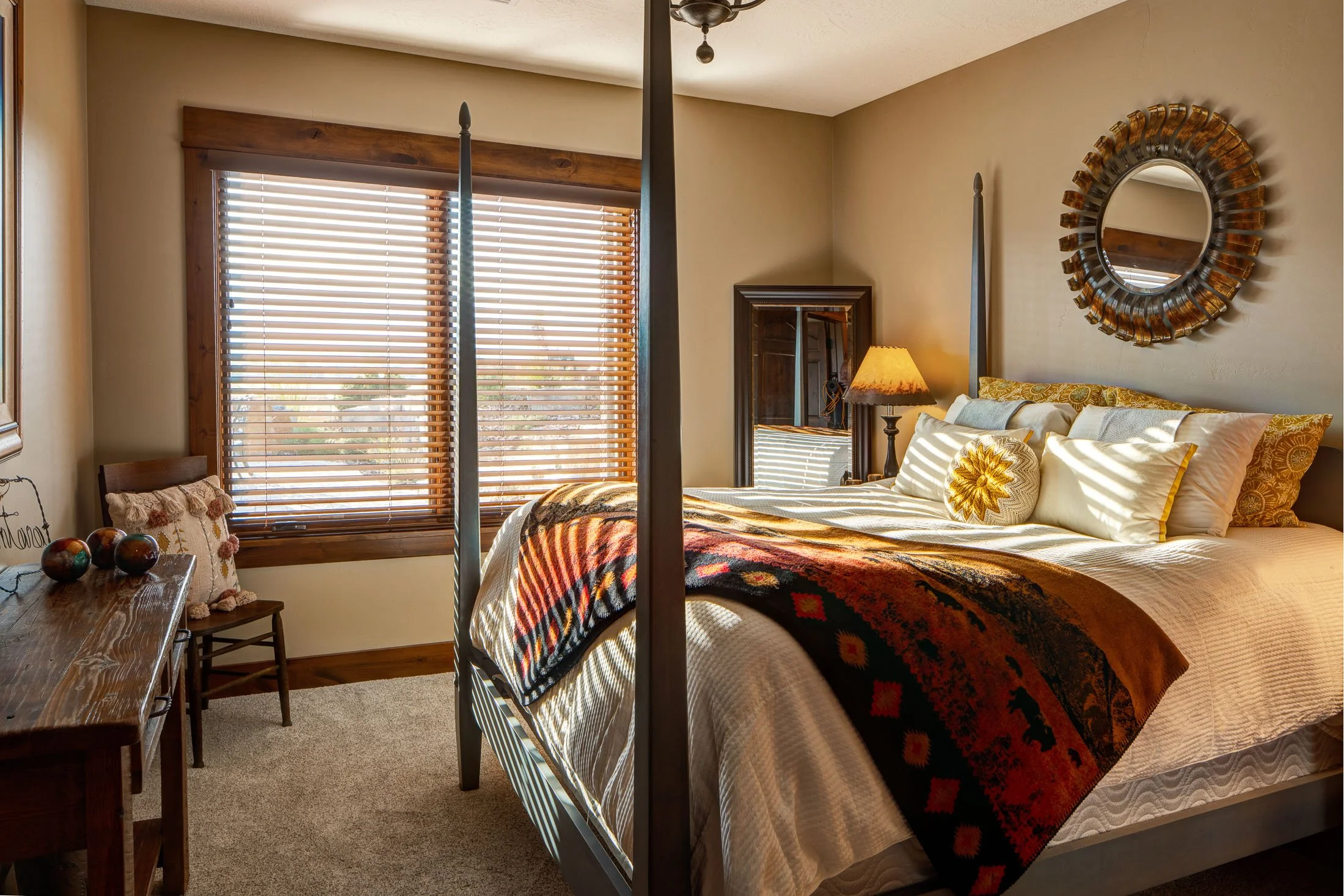 A cozy bedroom with a bed, four pillows, a textured blanket, a wooden nightstand with a lamp, a mirror on the wall, and a window with wooden blinds. Custom Home built in Montana by Edgell Building.