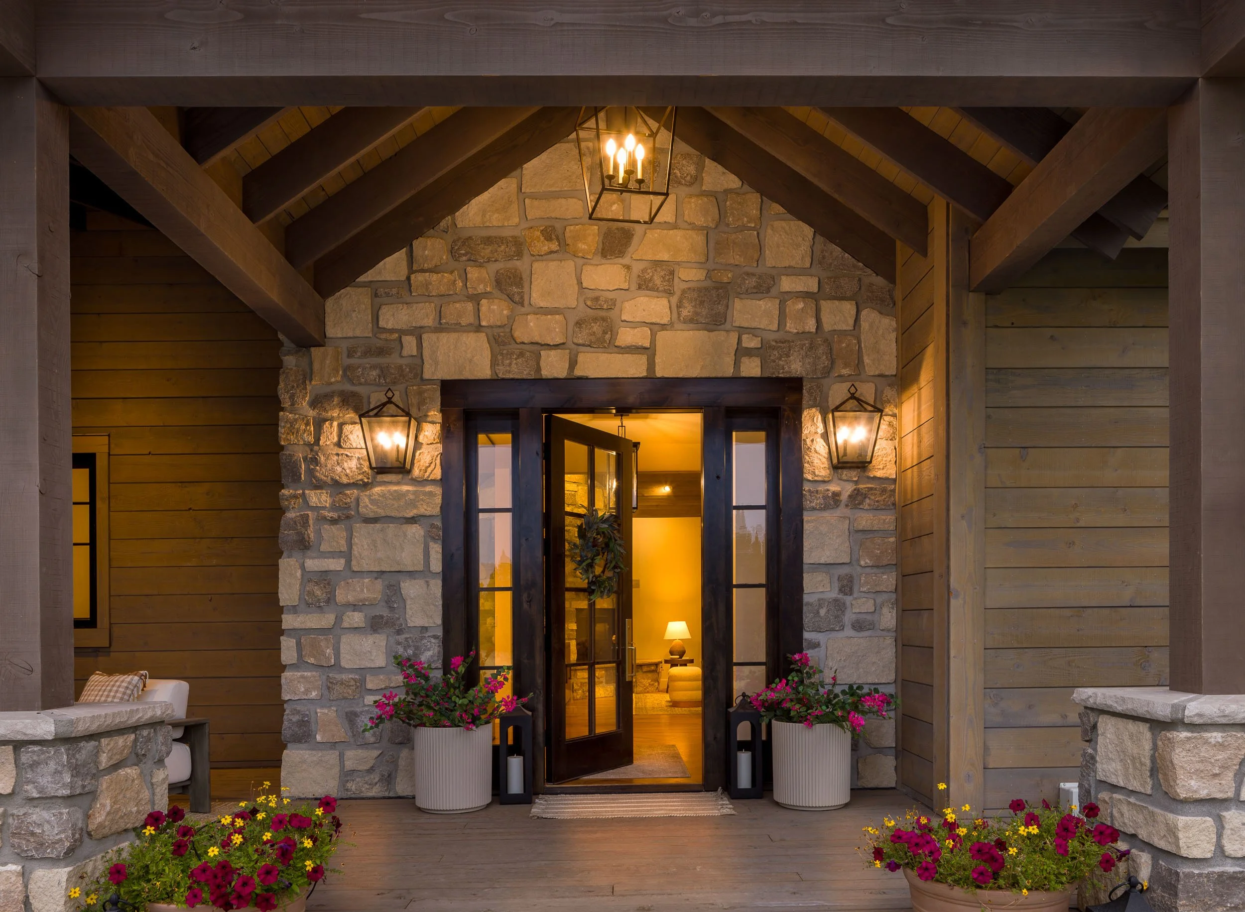 Front porch of a house with a stone and wood exterior, lit lanterns, potted flowers, and open glass door showing a lit interior with a lamp and furniture. Custom Home built in Montana by Edgell Building.