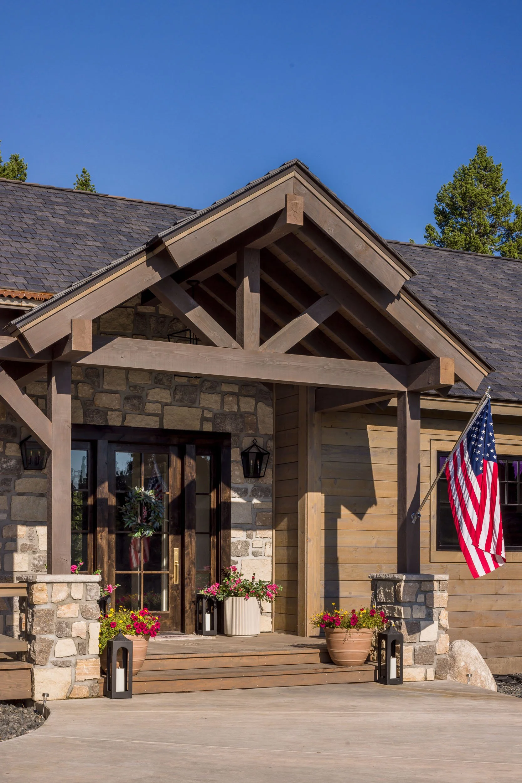 Front door of a house with stone and wood exterior, decorated with a wreath, potted flowers, and an American flag.