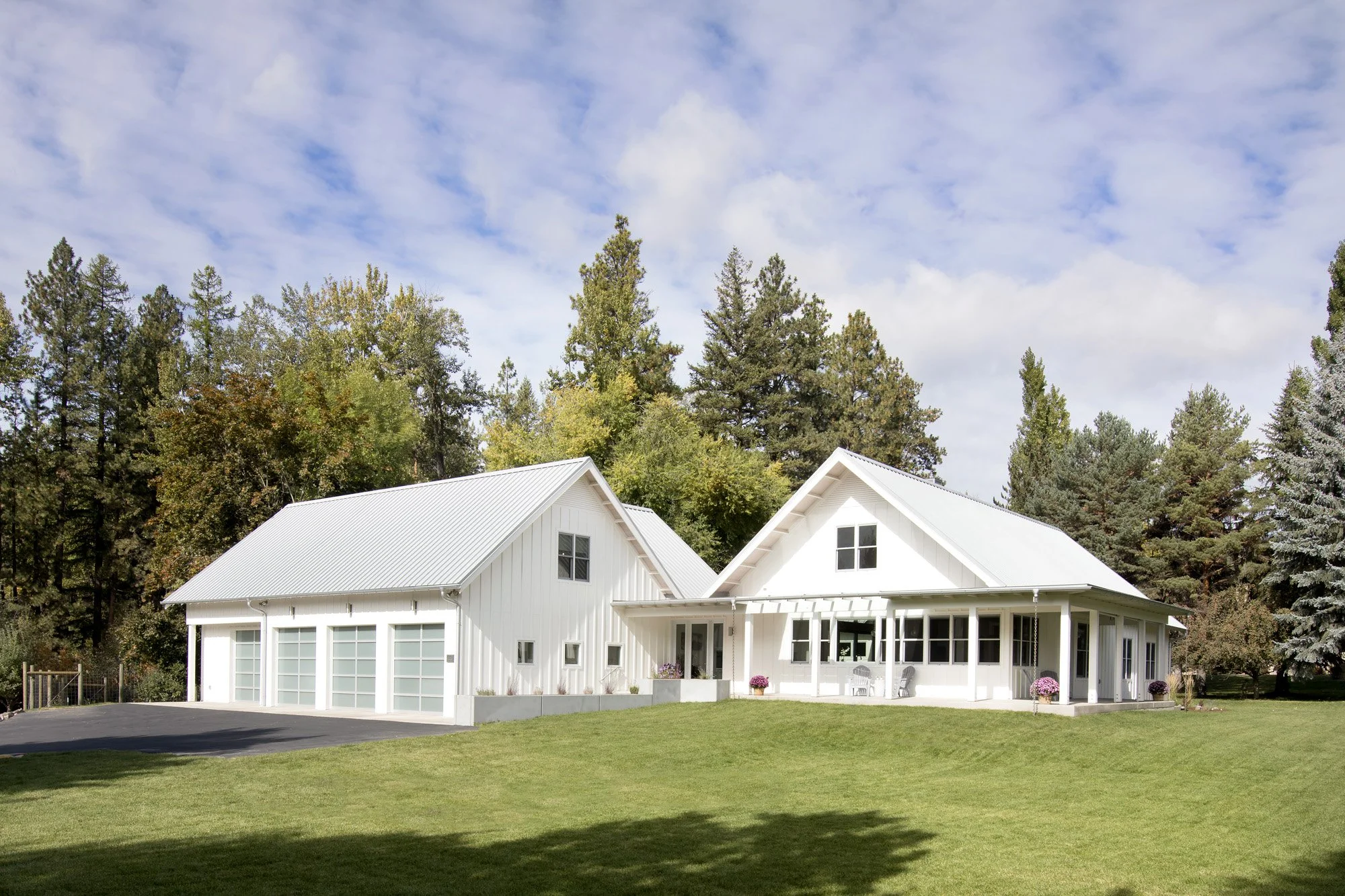 White house with a gray metal roof, surrounded by green grass and trees, under a partly cloudy sky. Custom Home built in Montana by Edgell Building.
