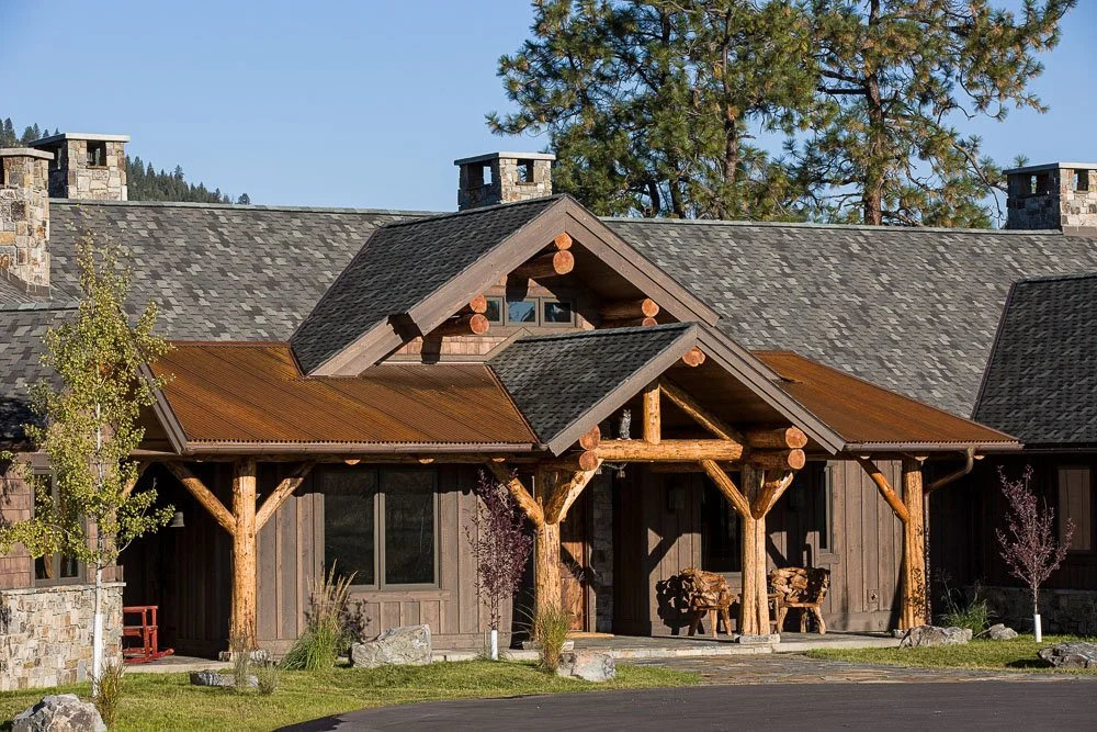 Log cabin-style house with wooden beams and stone accents, surrounded by trees and greenery, under a clear blue sky. Custom Home built in Montana by Edgell Building.