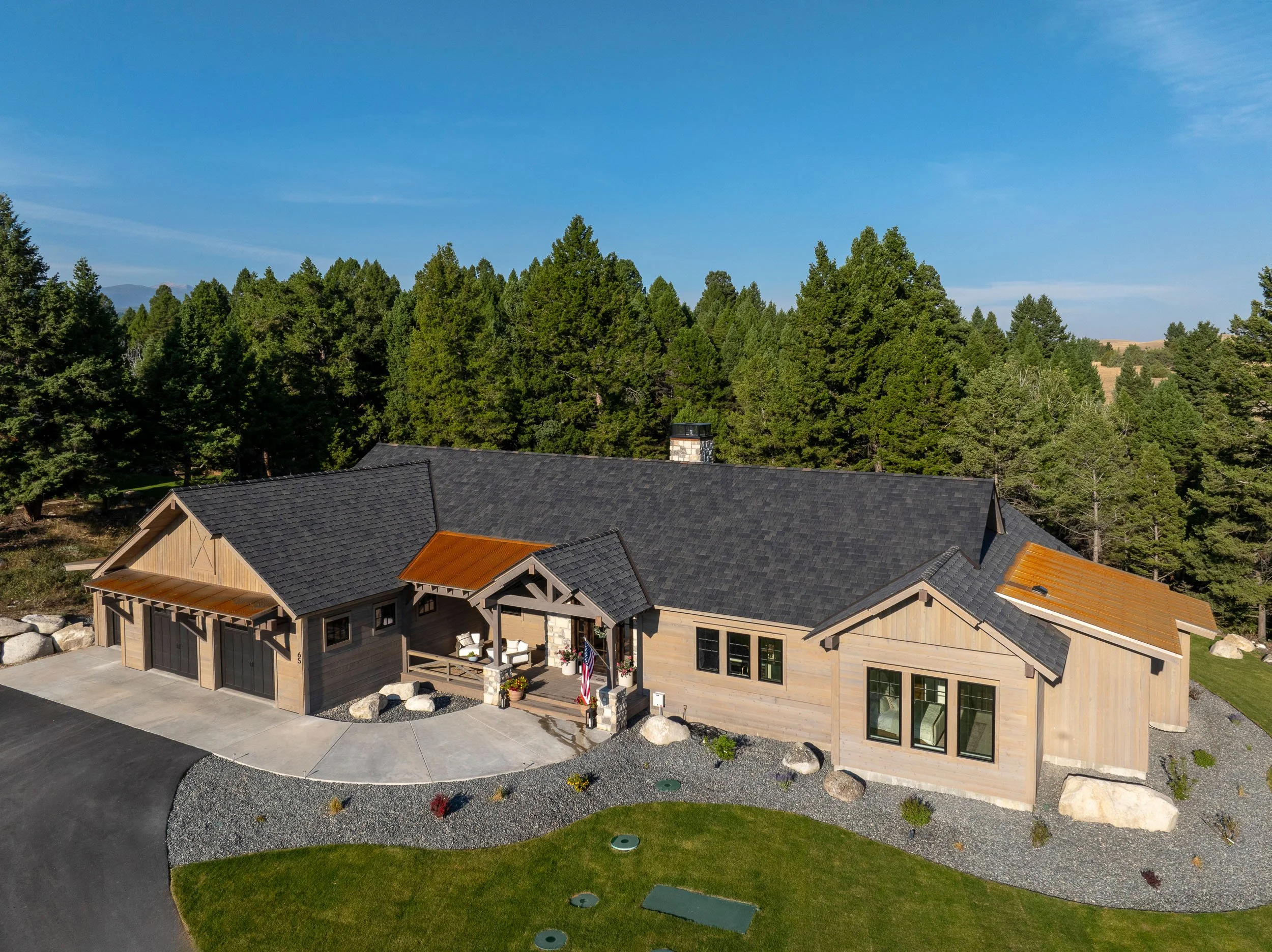 A modern house with a black roof, wooden exterior, and a front porch surrounded by trees and a landscaped yard. Home Designed and Built By Edgell Building in Western Montana.