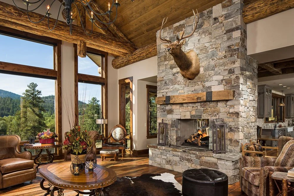 Living room with stone fireplace, mounted deer head, wooden beams, large windows showing mountain scenery, leather and upholstered furniture, chandelier, and decorative items. Custom Home built in Montana by Edgell Building.