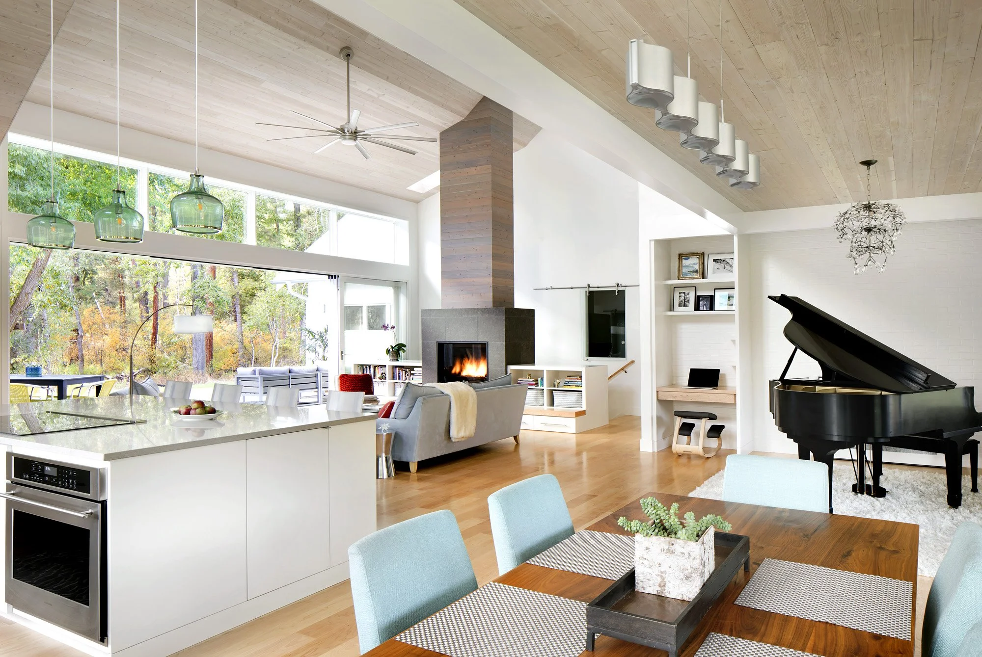 Open-concept living room with large windows, wood ceiling, hanging blue-green pendant lights, a fireplace, a black grand piano, a dining table with teal chairs, and a view of a wooded backyard. Home Designed and Built By Edgell Building in Montana.