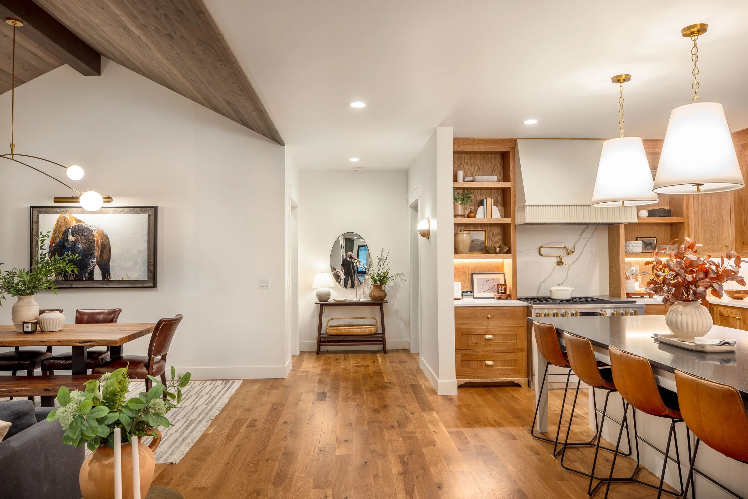 Open-plan kitchen and dining area with wood flooring, white walls, and modern decor. The kitchen features wooden cabinets, a marble backsplash, and a large island with barstools. Pendant lighting hangs above the island, and there are built-in shelves