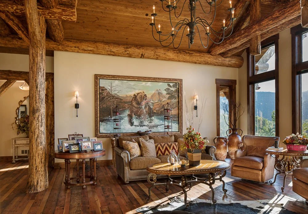 Rustic living room with large windows, wooden beams, and chandelier, decorated with framed photos, a large painting of a bear, and floral arrangements. Custom Home built in Montana by Edgell Building.