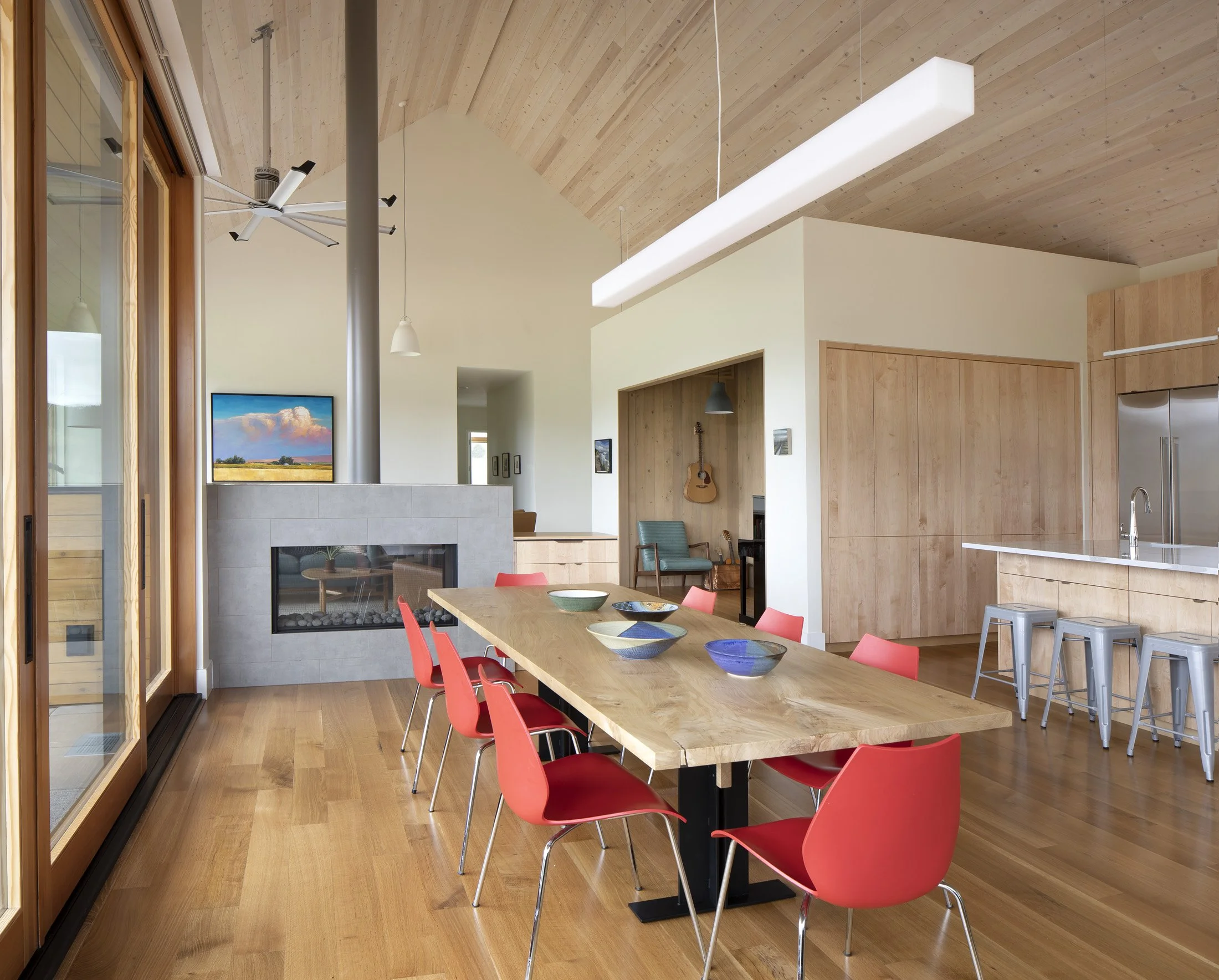 Modern open-concept dining area with wooden floors, a large wooden dining table with red chairs, and a kitchen with a bar and stools. Features include a fireplace, a wall-mounted TV. Custom Home built in Montana by Edgell Building.