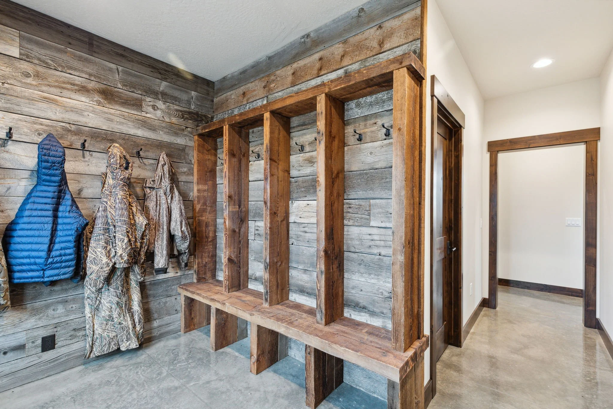 Mudroom with wooden wall panels, hooks for hanging jackets, a wooden coat rack with cubbies, and camouflage outdoor clothing. Custom Home built in Montana by Edgell Building.