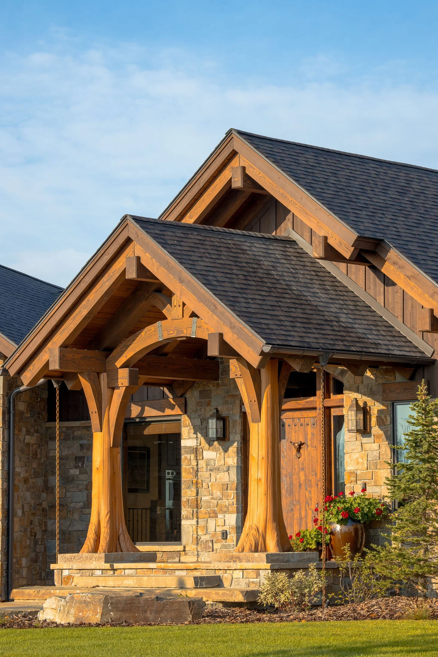 Front view of a house with a stone exterior, wooden accents, and a porch with large wooden columns, decorated with a pot of red flowers, under a blue sky. Custom Home built in Montana by Edgell Building.