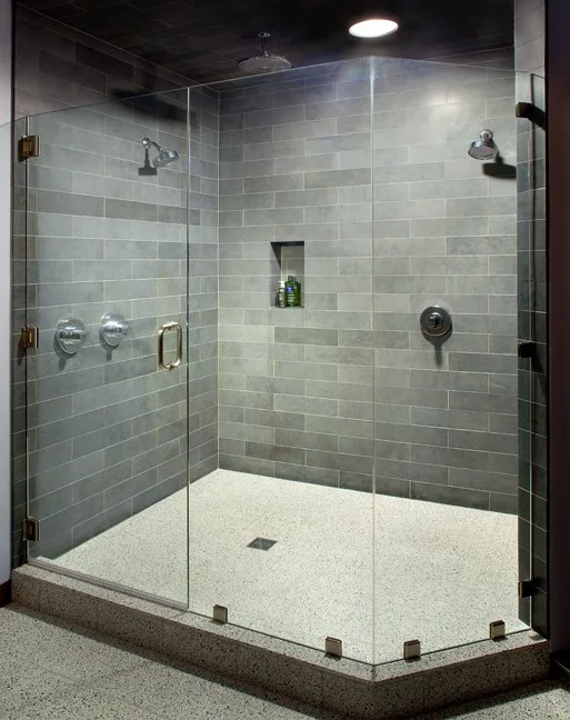 A modern walk-in shower with glass doors, grey tiled walls, and a pebble-textured floor. It has two showerheads and a built-in niche with toiletries. Custom Home built in Montana by Edgell Building.