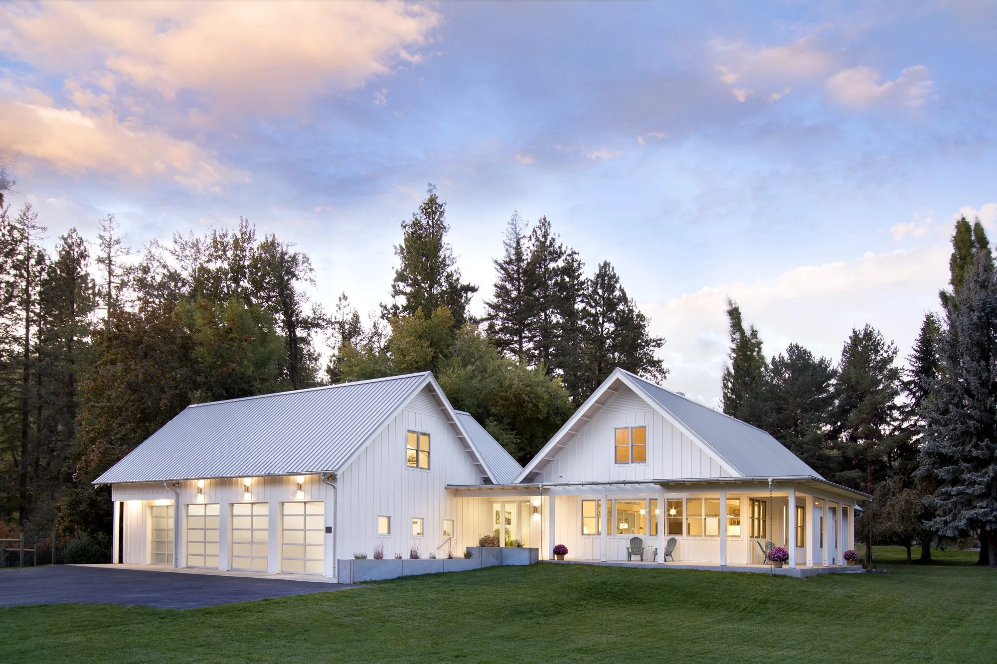 A modern white house with a metal roof and large windows, illuminated from inside, surrounded by a grassy yard and tall trees, during dusk with a partly cloudy sky. Custom Home built in Montana by Edgell Building.