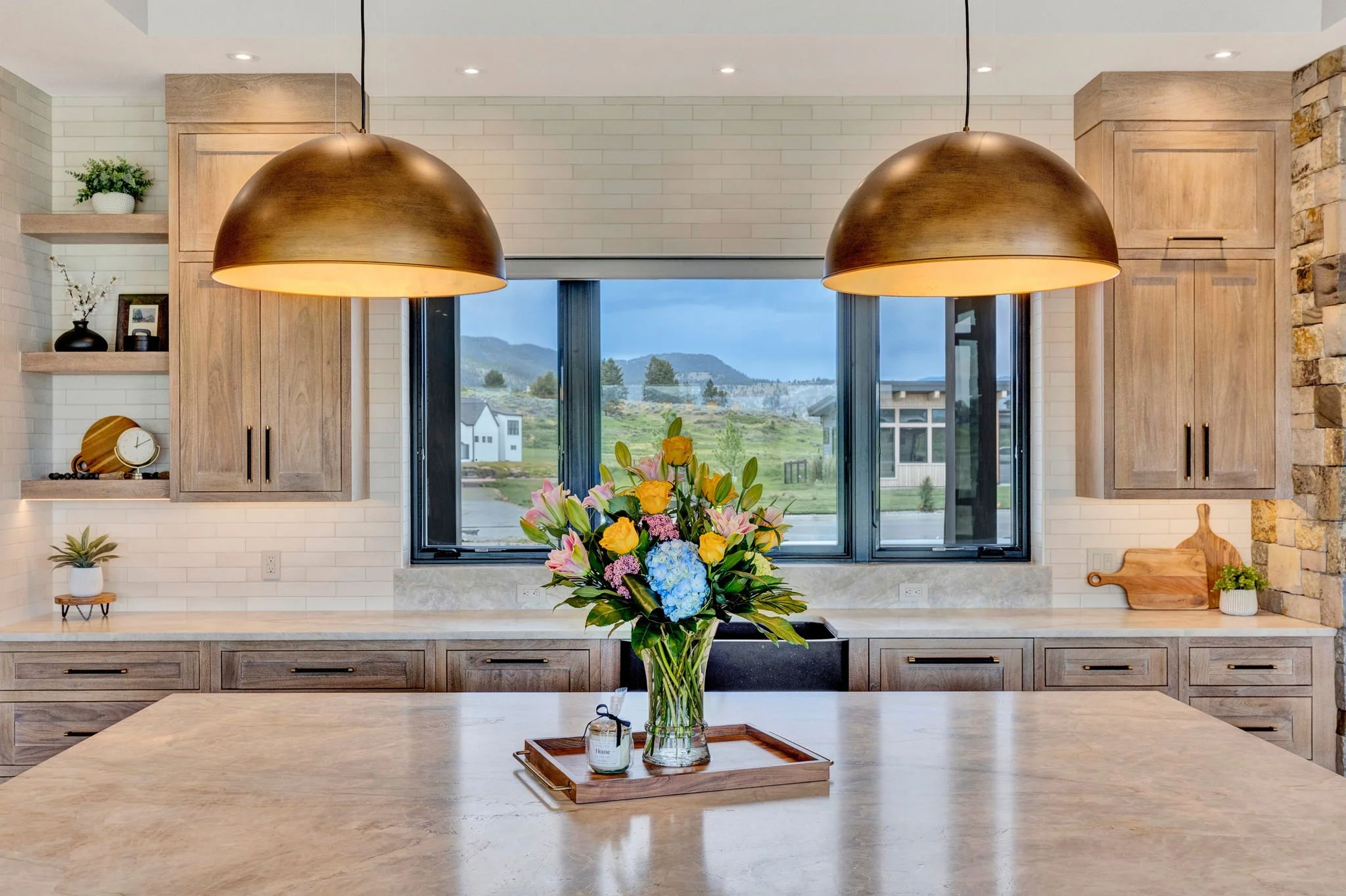 Modern kitchen with light wood cabinets, two large pendant lights, a window showing a scenic outdoor landscape, a flower arrangement on the countertop, and shelves with decorative items. Custom Home built in Montana by Edgell Building.