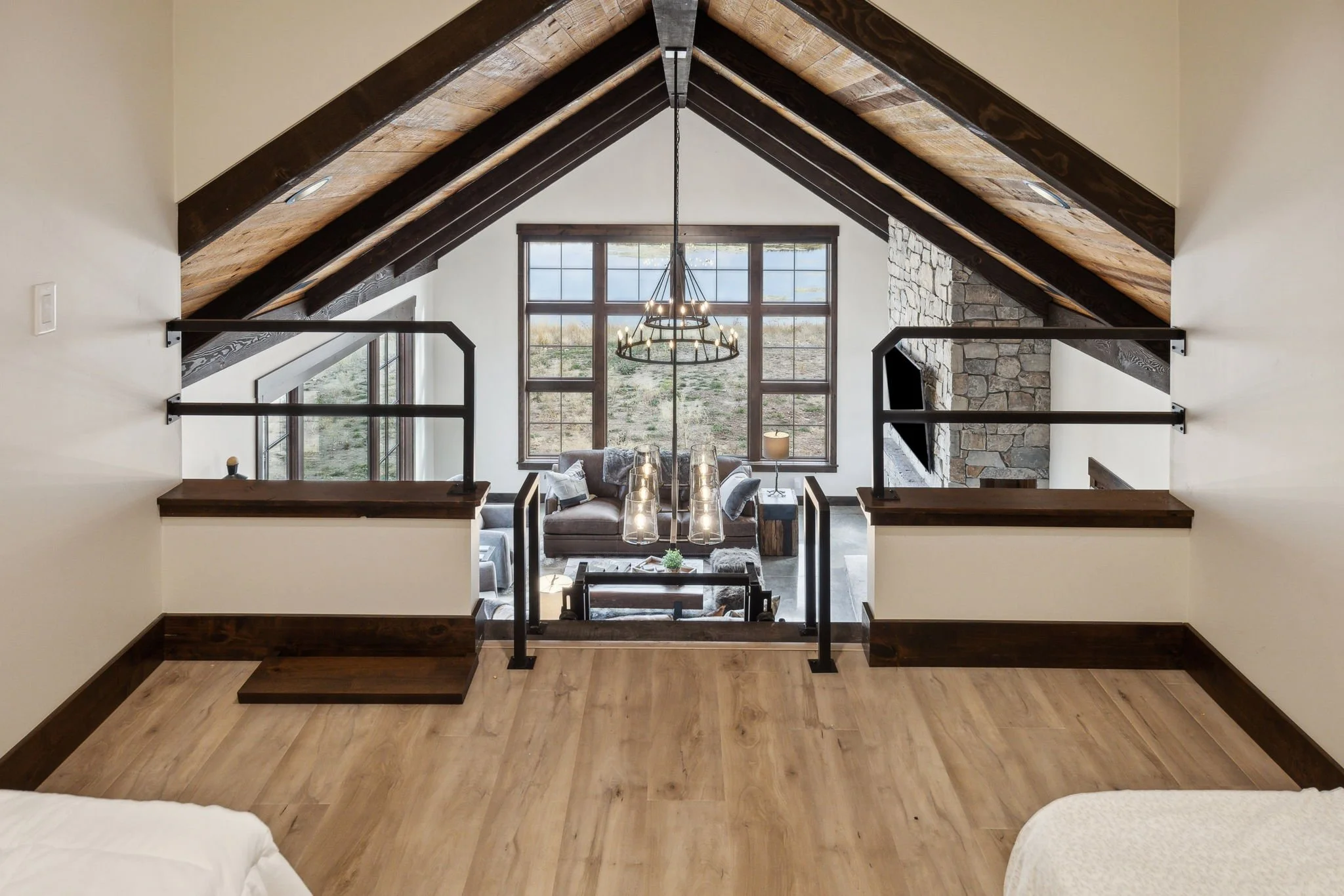 View of a living room with large windows, stone fireplace, modern chandelier, sofa, table, and decorative pillows, through an open upper level with black metal railings. Custom Home built in Montana by Edgell Building.