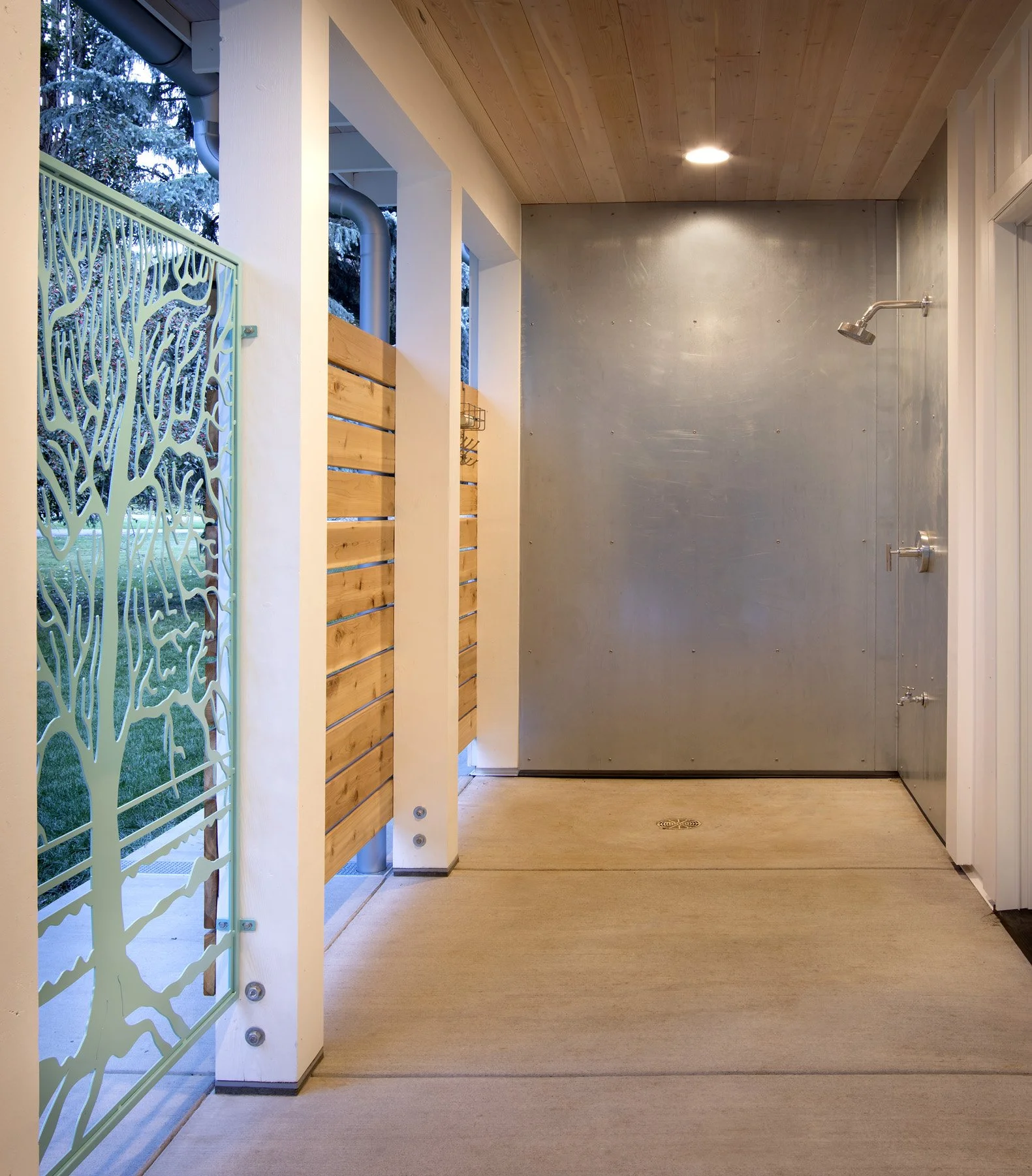 An outdoor shower area with a concrete wall, a shower head, and a drain in the floor, partially enclosed by a wooden privacy screen and a decorative metal gate with a tree design. Custom Home built in Montana by Edgell Building.