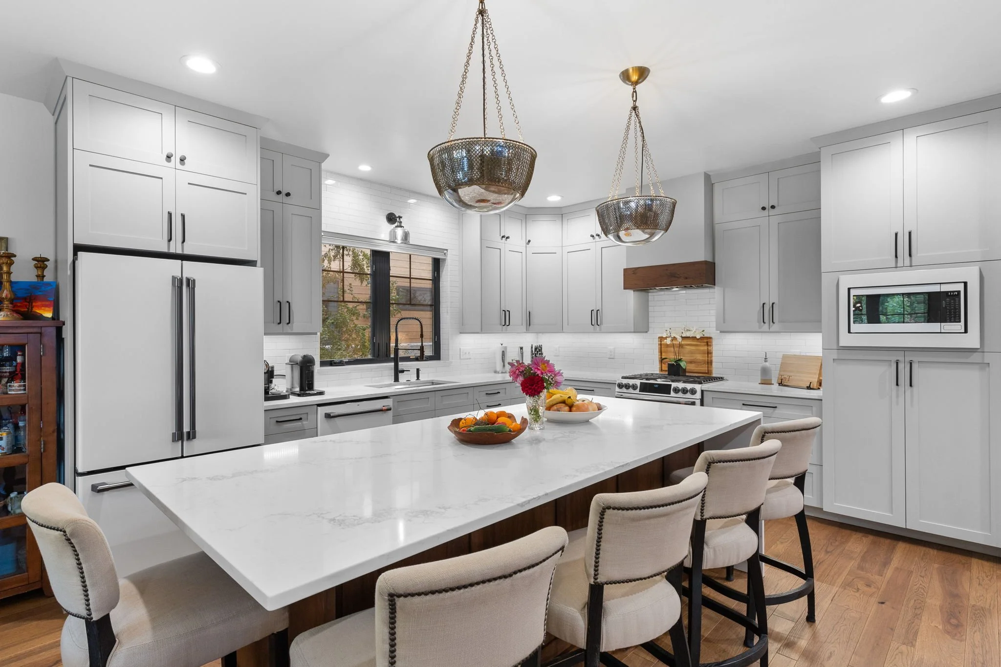 Modern kitchen with white cabinets, a large island with a white marble top, beige chairs, fruit bowls, pendant and recessed lighting, black window, white brick backsplash, and stainless steel appliances. Custom Home built in Montana by Edgell Buildin