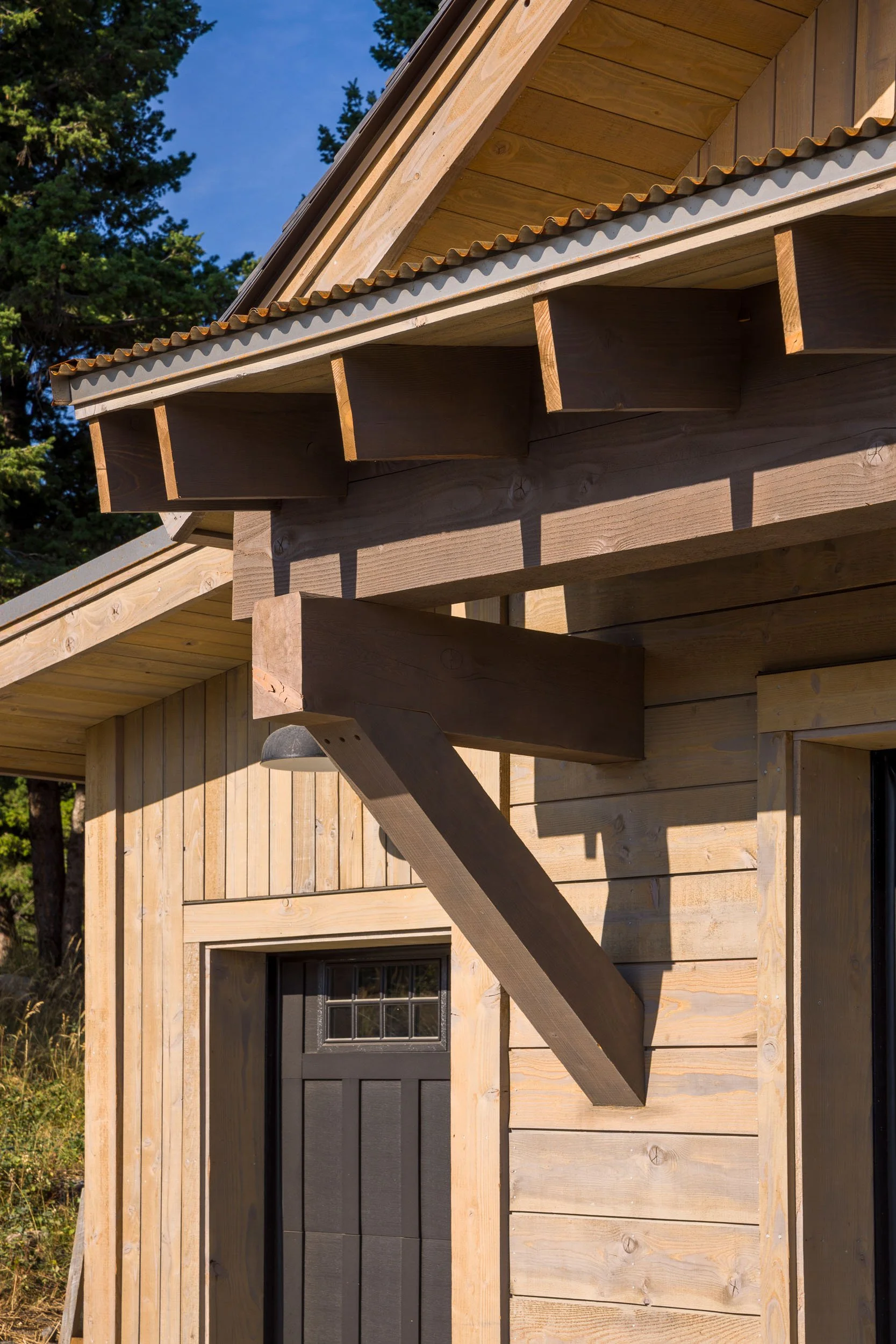 Close-up of a wooden house with exposed structural elements, including beams and siding, with a garage door below and trees in the background. Custom Home built in Montana by Edgell Building.