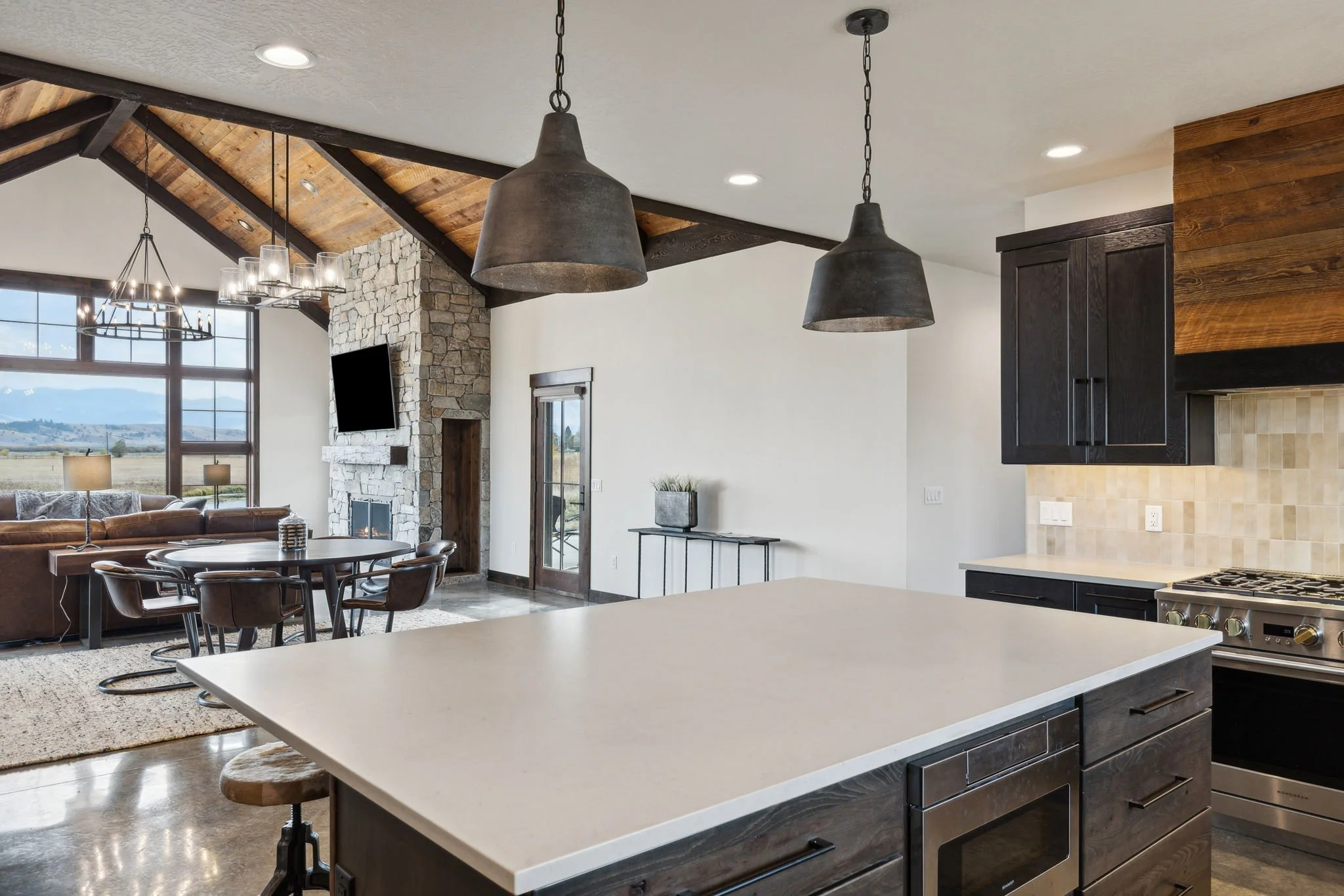 Modern open-concept kitchen and living room with high vaulted ceiling, stone fireplace, large window with view, round dining table with chairs, and hanging pendant lights. Custom Home built in Montana by Edgell Building.