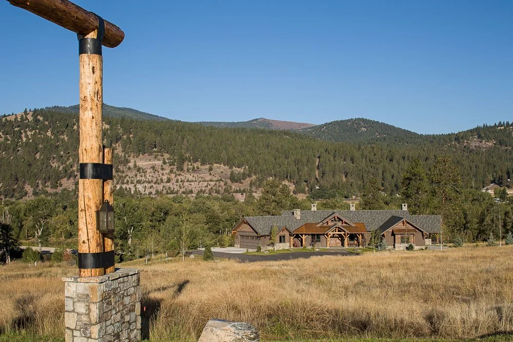 A wooden archway with mounted lantern in the foreground, a large house with multiple sections and a grey roof in the middle ground, and forested mountains under a clear blue sky in the background. Custom Home built in Montana by Edgell Building.
