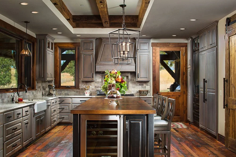 Rustic kitchen with gray cabinets, a large wooden island, a wine fridge, and wooden beams on the ceiling. Window views of the outdoors. Custom Home built in Montana by Edgell Building.