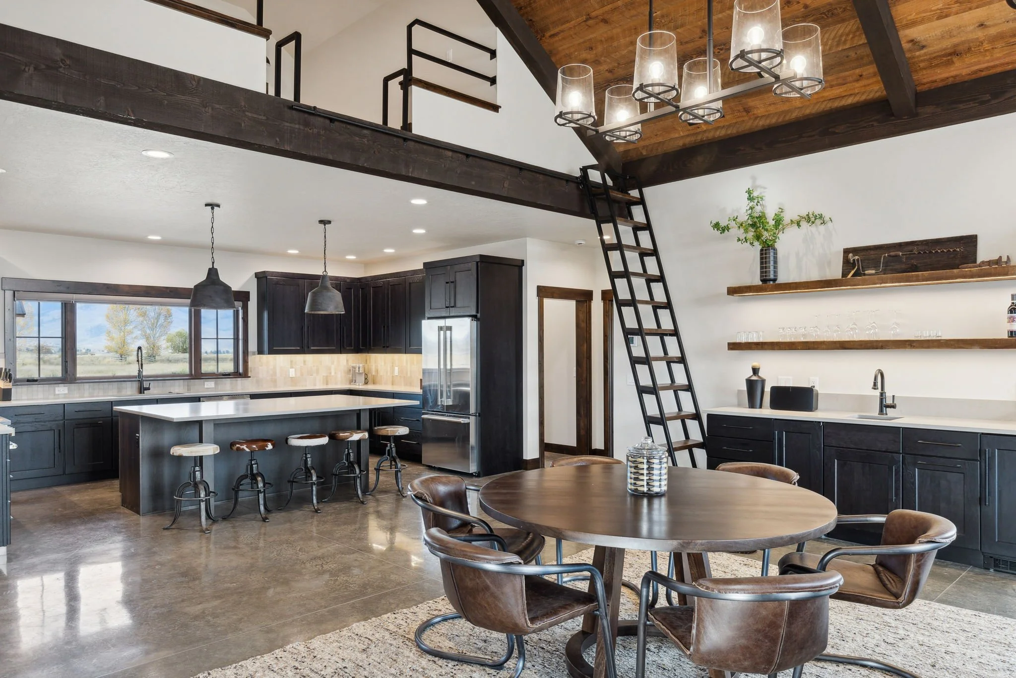 Open-concept kitchen and dining area with dark cabinets, white countertops, a kitchen island with bar stools, a round dining table with leather chairs, and a loft with a ladder and chandelier. Custom Home built in Montana by Edgell Building.