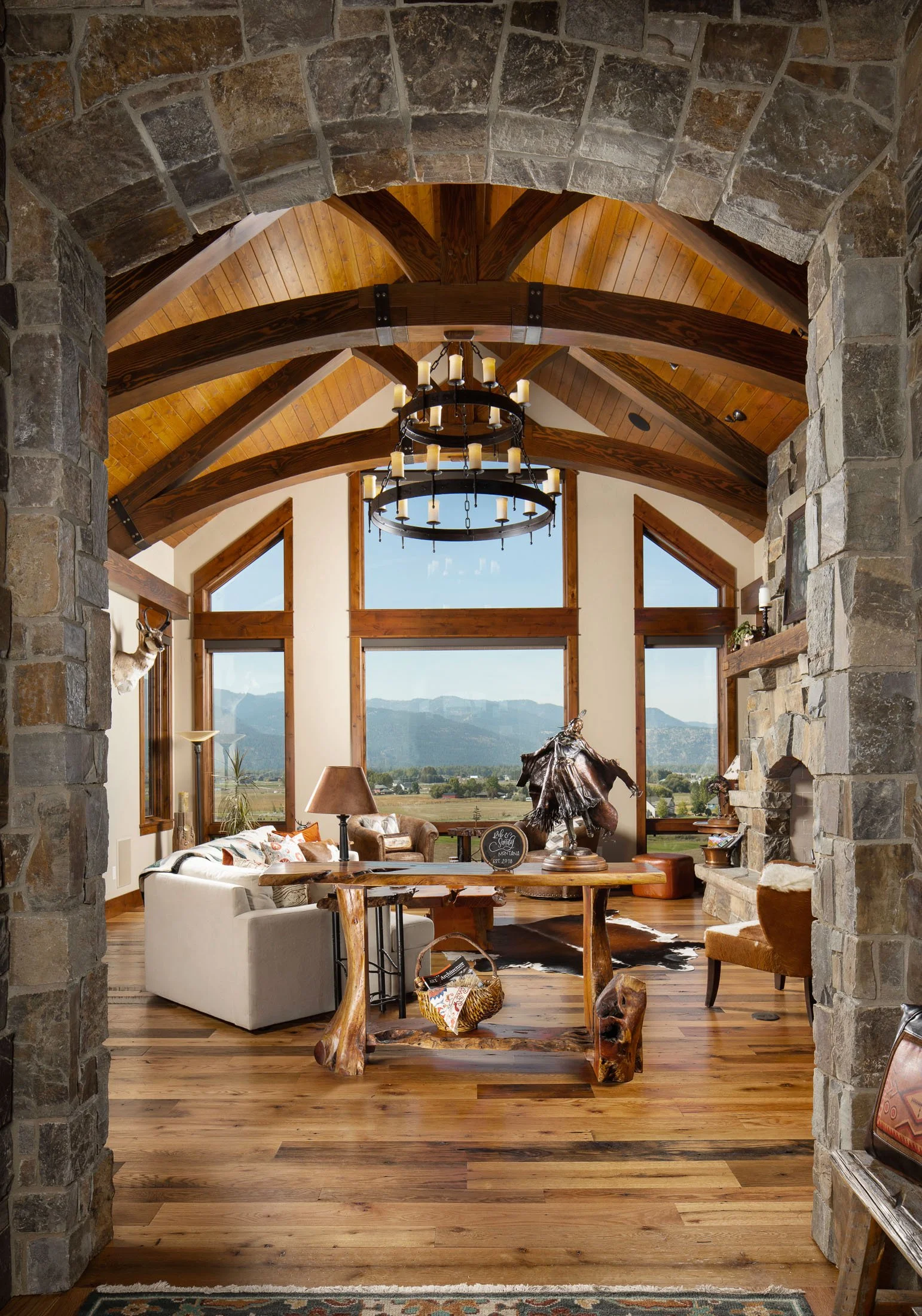 A living room with a rustic style featuring large stone fireplace, wooden vaulted ceiling with beams, large windows displaying mountain and blue sky view, and log furniture including a table and chairs. Custom Home built in Montana by Edgell Building