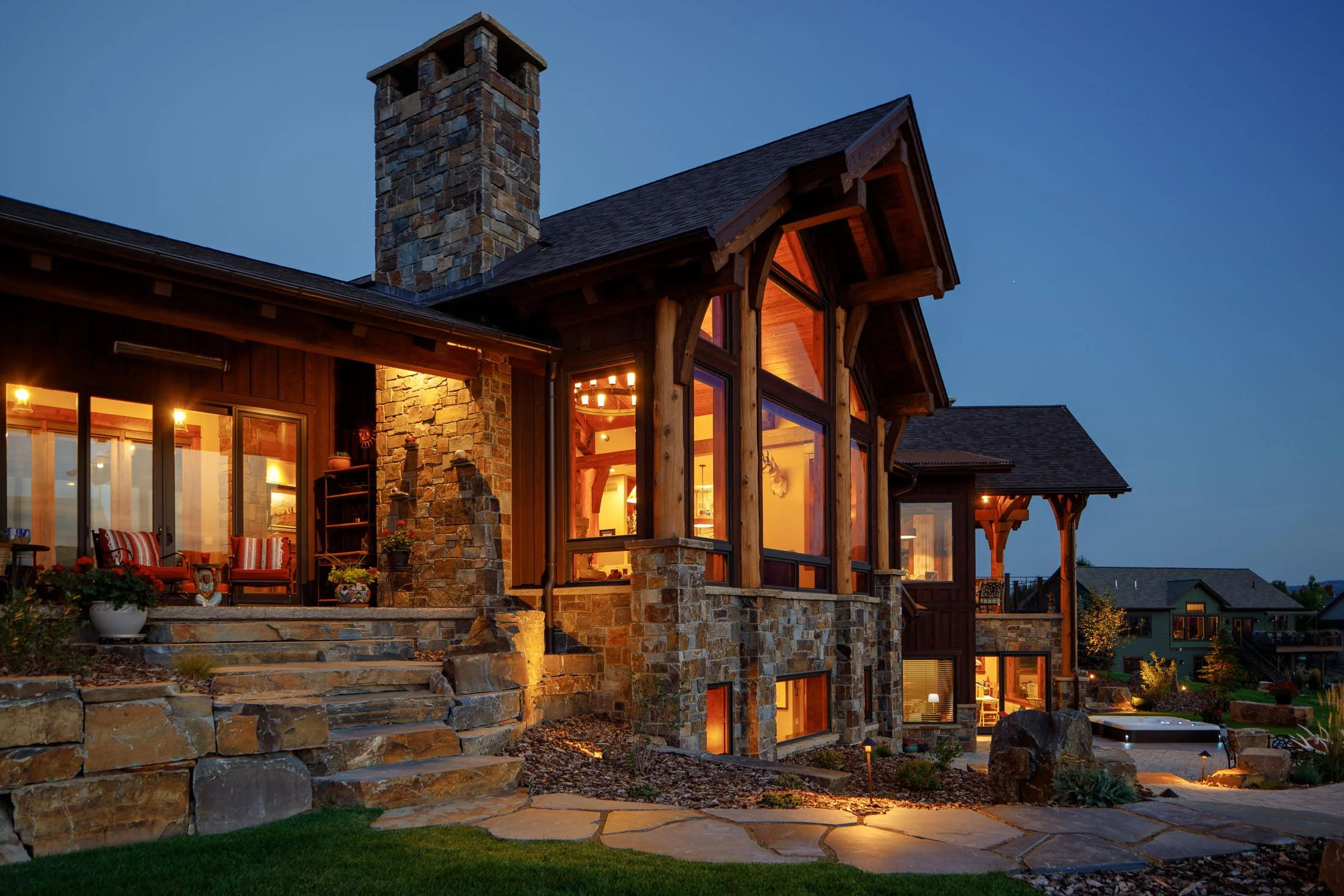 A house at dusk with large windows, stone and wood exterior, front steps, and a well-lit interior visible through the windows. Custom Home built in Montana by Edgell Building.